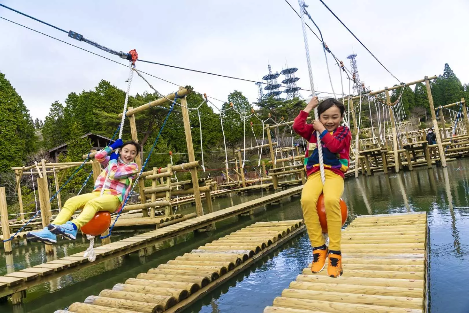 兩位孩童在日本六甲GREENIA運動公園的水上滑索設施上開心地玩耍，體驗刺激的空中運動樂趣，背景綠意盎然。