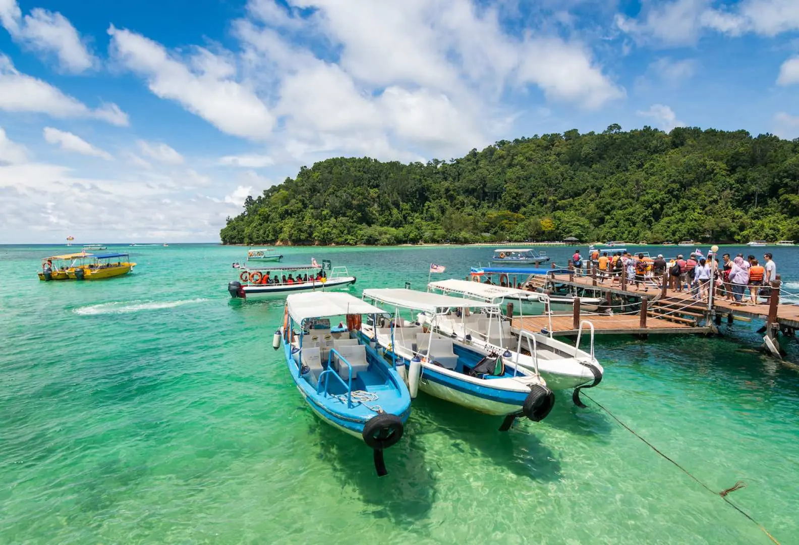 Boats docked at a scenic wooden jetty leading to a lush green tropical island with turquoise waters, offering a perfect spot to relax between dives in Sabah's marine park.