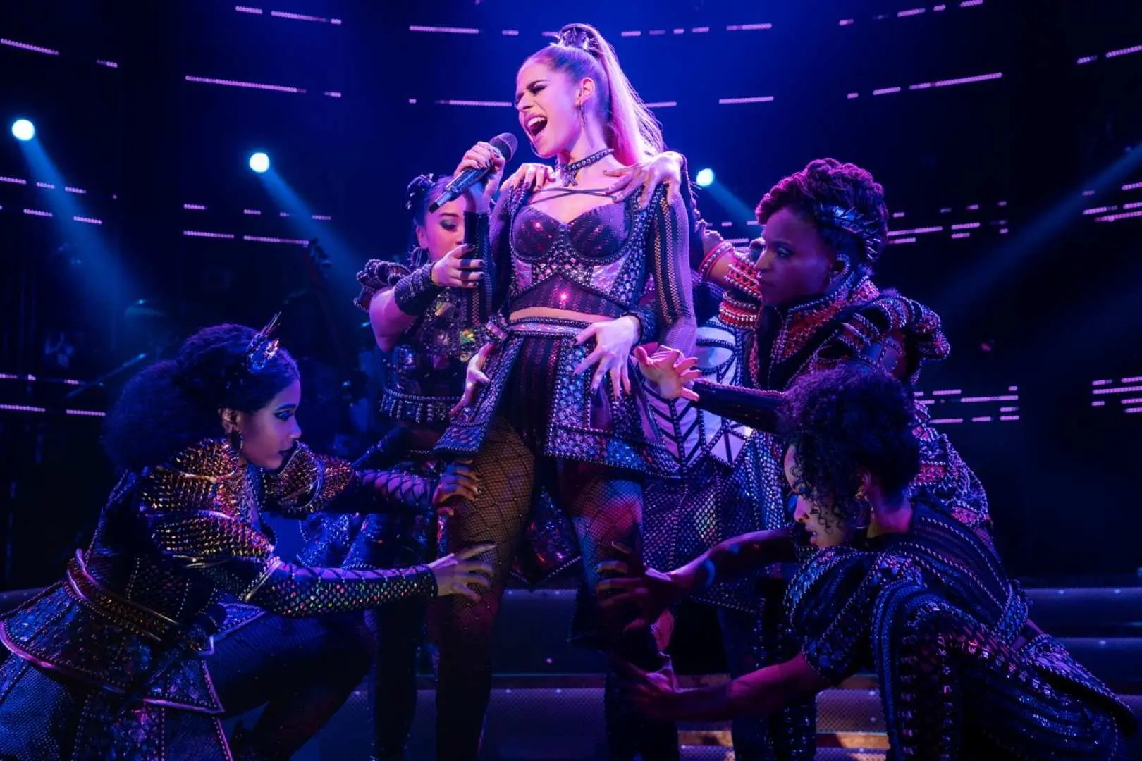 A central performer in the New York Broadway musical SIX singing soulfully under blue stage lights, with other cast members reaching out, creating a dramatic and emotionally charged scene.