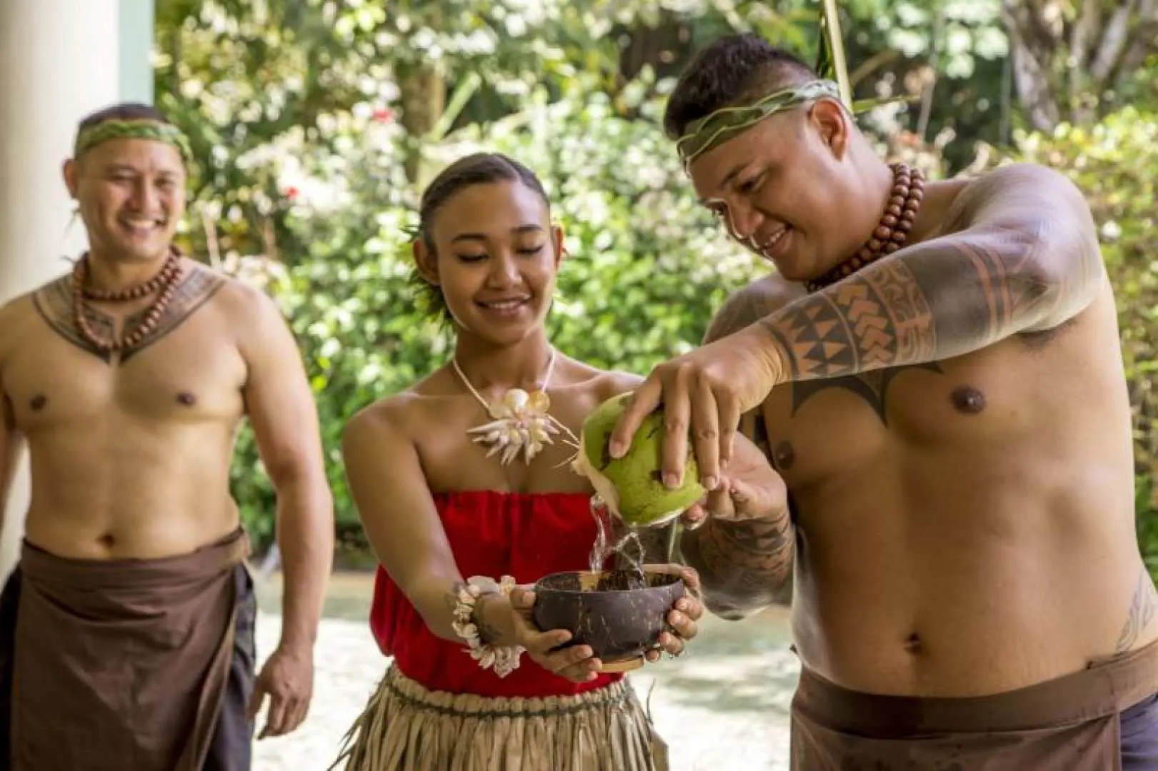 Visitors in traditional costumes taste fresh coconut water at Guam's Fisheye Marine Park, as islanders demonstrate coconut milk extraction, immersing in vibrant Pacific island culture.