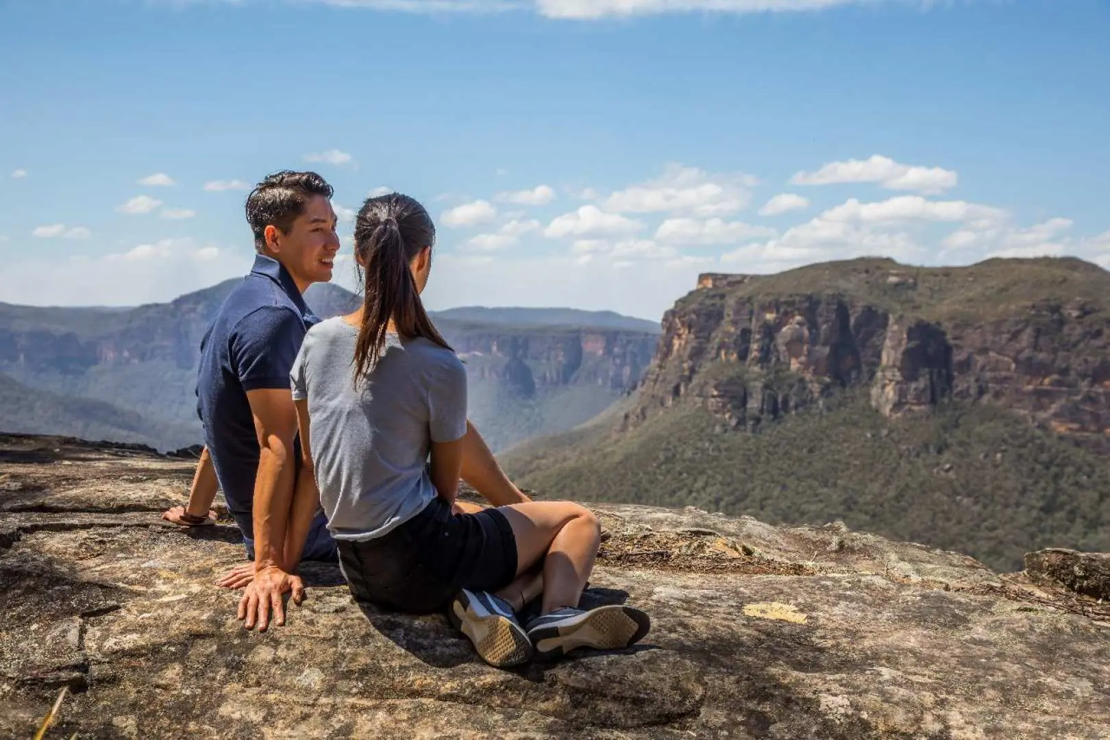 Travelers relaxing on a cliff edge in Blue Mountains, gazing at the expansive Jamison Valley and rugged ranges under a clear afternoon sky.