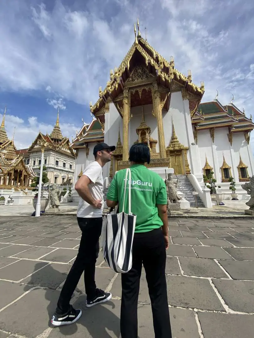 A tourist and 'TripGuru' guide walk towards a golden-roofed Thai building within Bangkok's Grand Palace. They are exploring Thailand's royal residence and must-see landmarks.