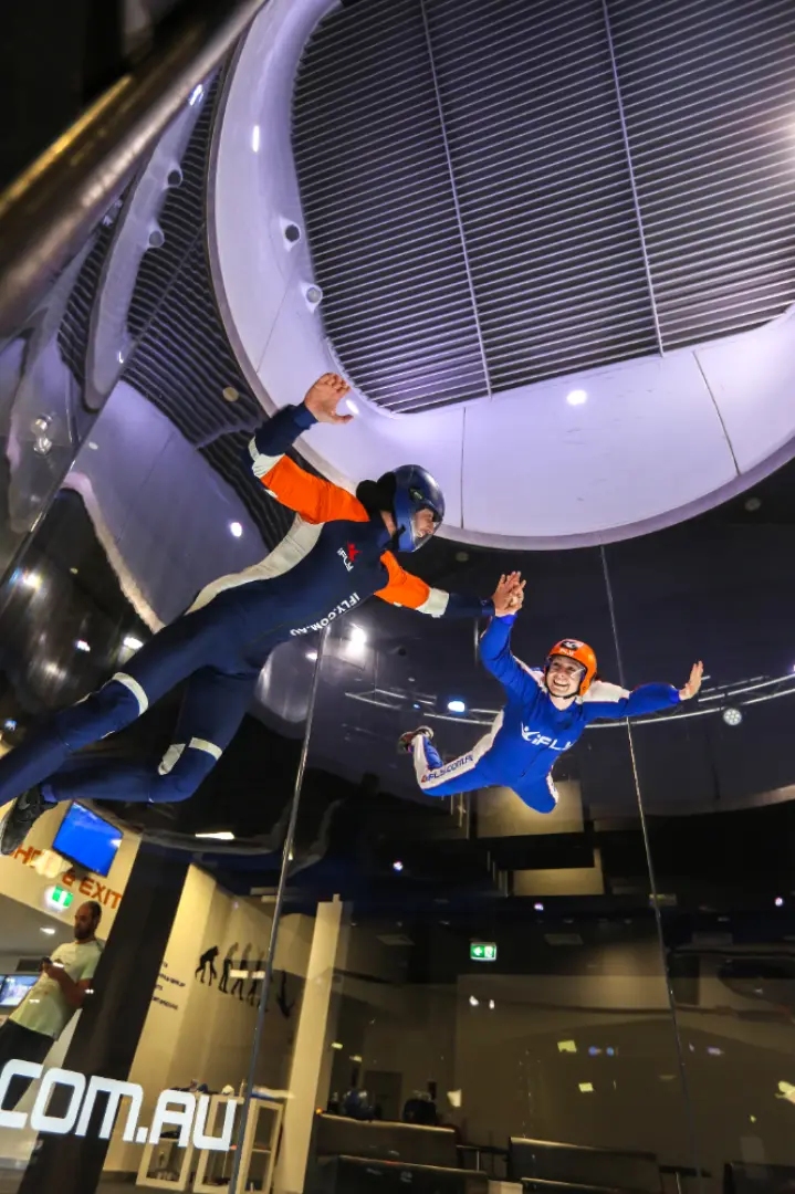 Gold Coast iFLY indoor skydiving: A smaller participant, possibly a child, joyfully soars in the transparent wind tunnel, receiving careful one-on-one guidance from an instructor for a safe and exhilarating flight.