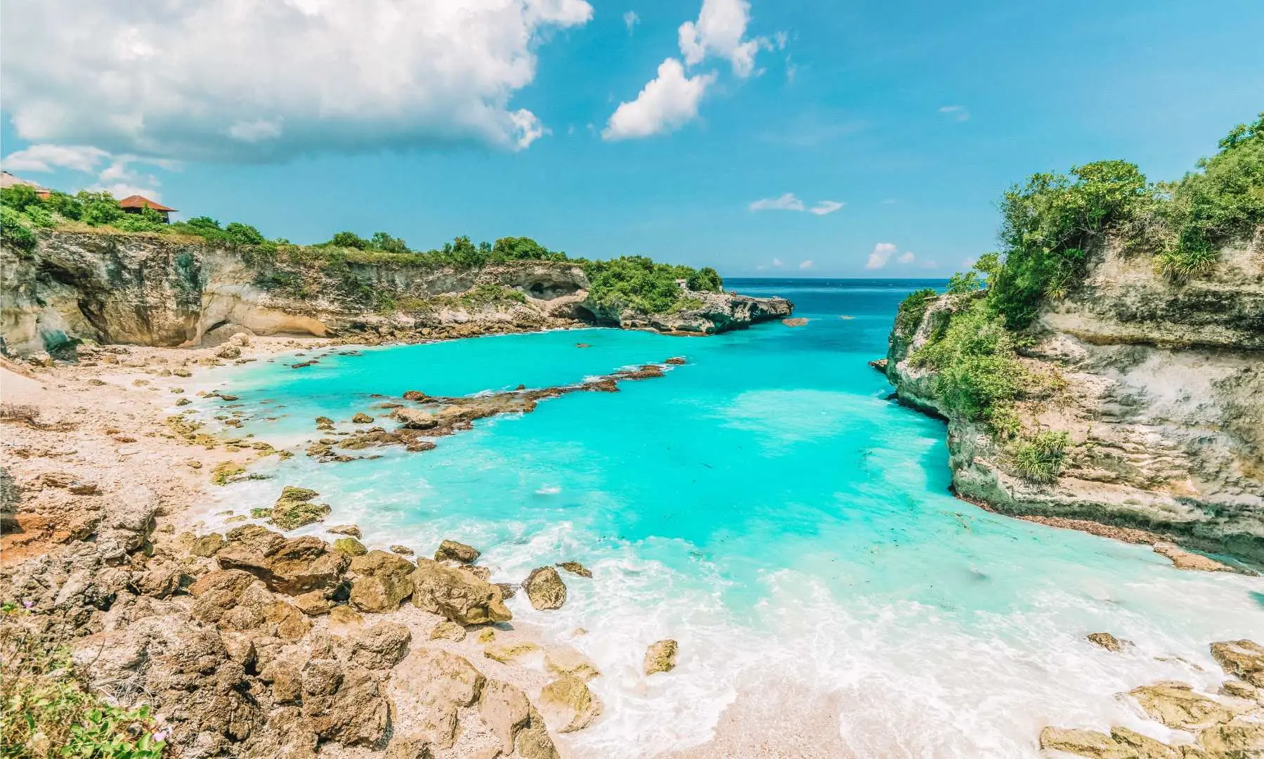 Stunning panoramic view of Bali's Tanjung Jepun, showcasing a secluded turquoise cove with white sand and rock formations, surrounded by lush green cliffs under a clear sky.