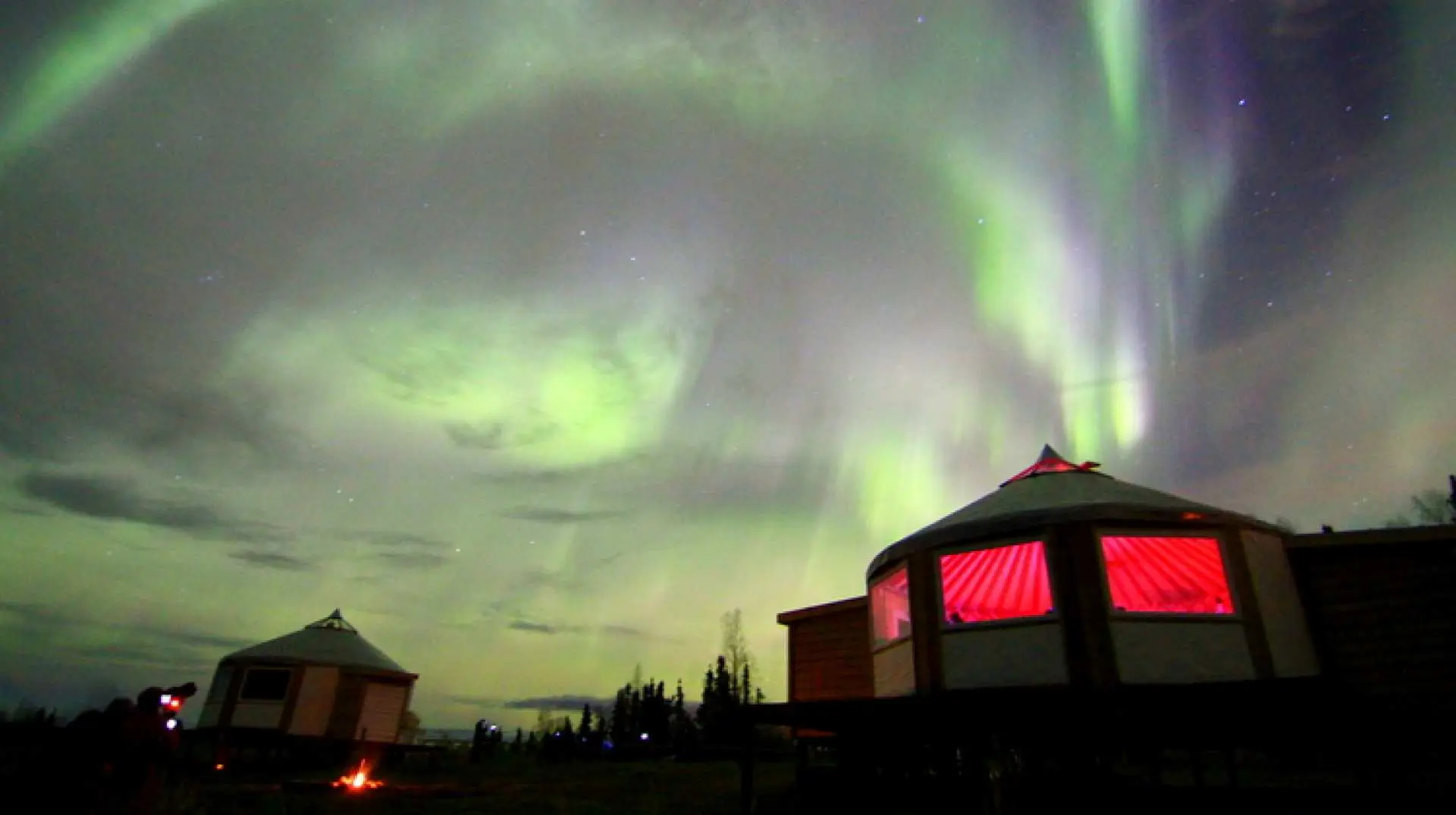 Alaskan aurora cabins under a vibrant green aurora, with one cabin's windows glowing red, offering a warm indoor and excellent outdoor viewing experience near Fairbanks.