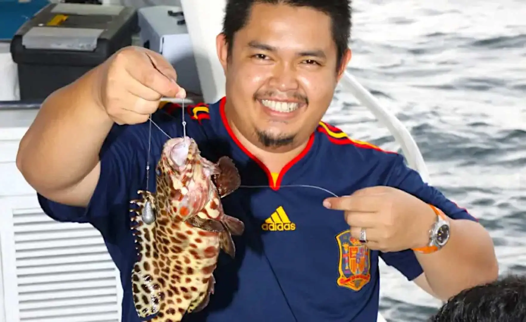 Smiling tourist on a boat proudly holding a freshly caught spotted grouper fish during a boat fishing experience at Usukan Cove and Pandan Pandan Island, Sabah.