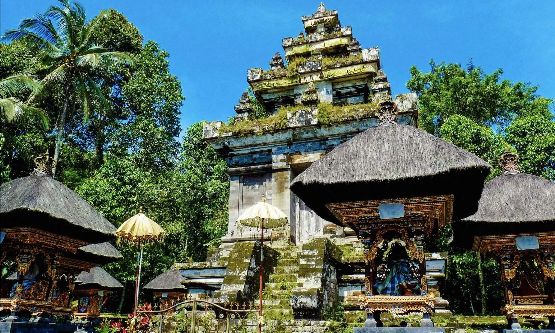 The majestic stone architecture of Bali's Pura Menninging Water Temple, with its multi-tiered roofs under a blue sky, surrounded by lush tropical vegetation.