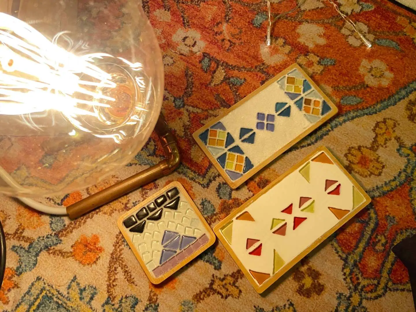 Three handcrafted mosaic coasters in various geometric patterns and vibrant colors displayed on a patterned rug next to a vintage light bulb, showcasing personalized artwork from the DIY workshop.
