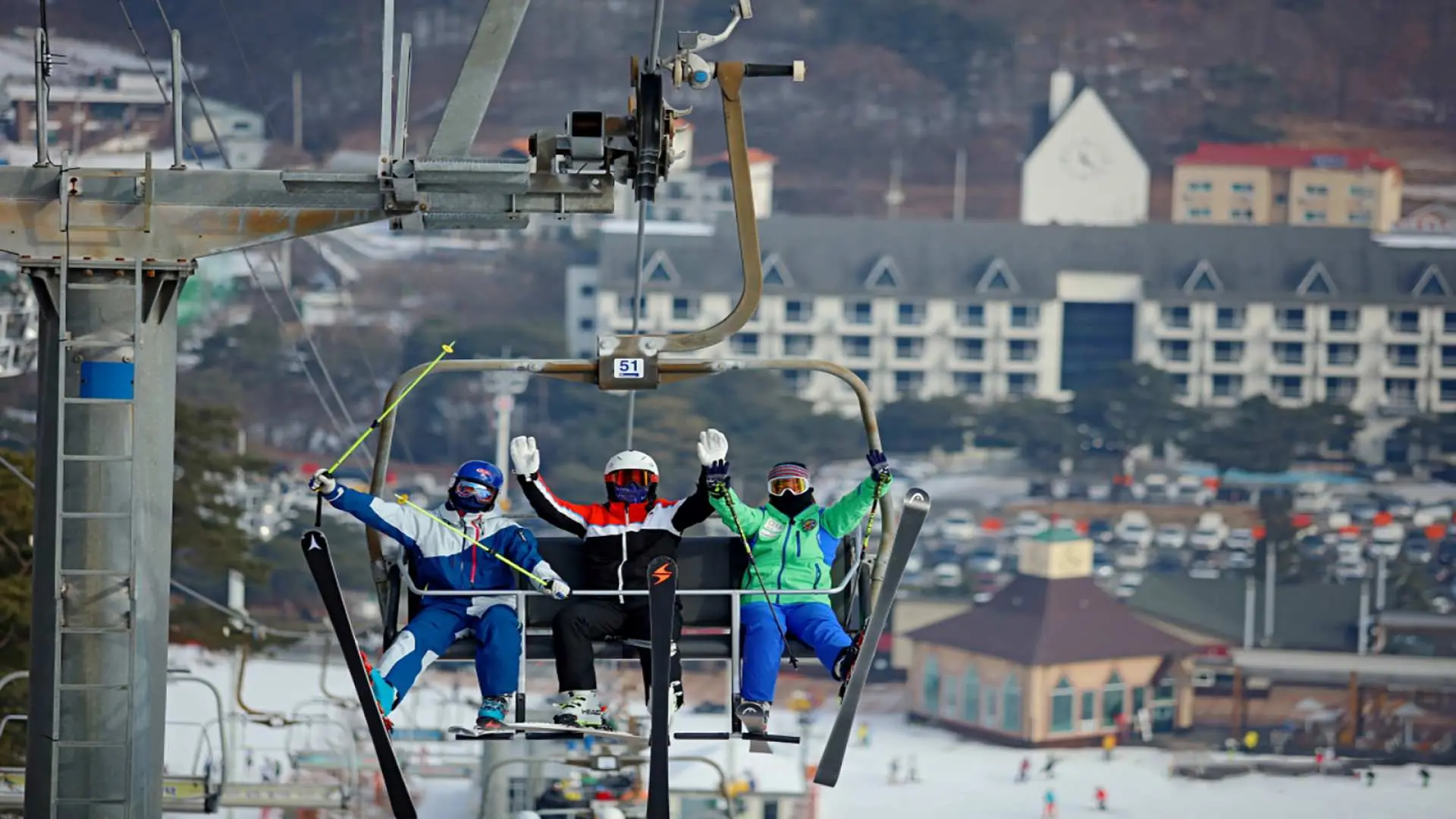 Three skiers excitedly waving from a chairlift ascending the slopes of Jisan Ski Resort in South Korea, offering a view of the buildings and parking lot below, ready for thrilling ski adventures.