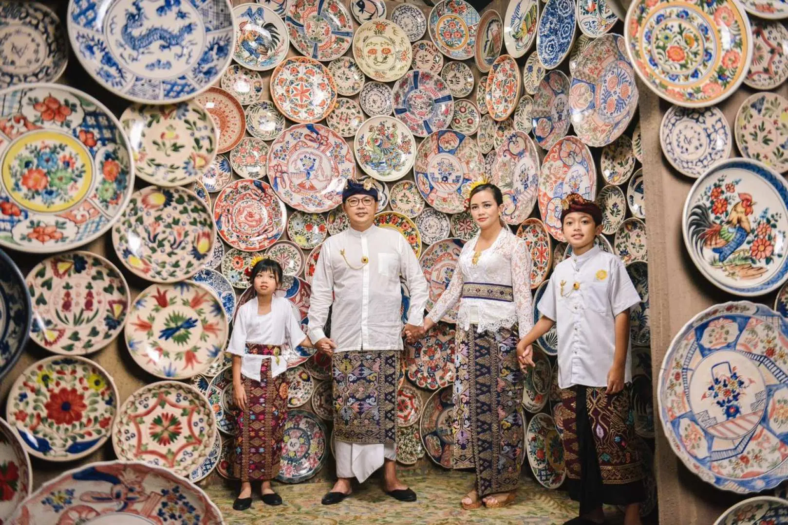 ครอบครัวในชุดบาหลีพื้นเมืองกำลังยิ้มหน้ากำแพงที่ตกแต่งอย่างประณีตด้วยจานเซรามิกหลากสีสัน ที่ Asana Artseum ในสวนวัฒนธรรม GWK