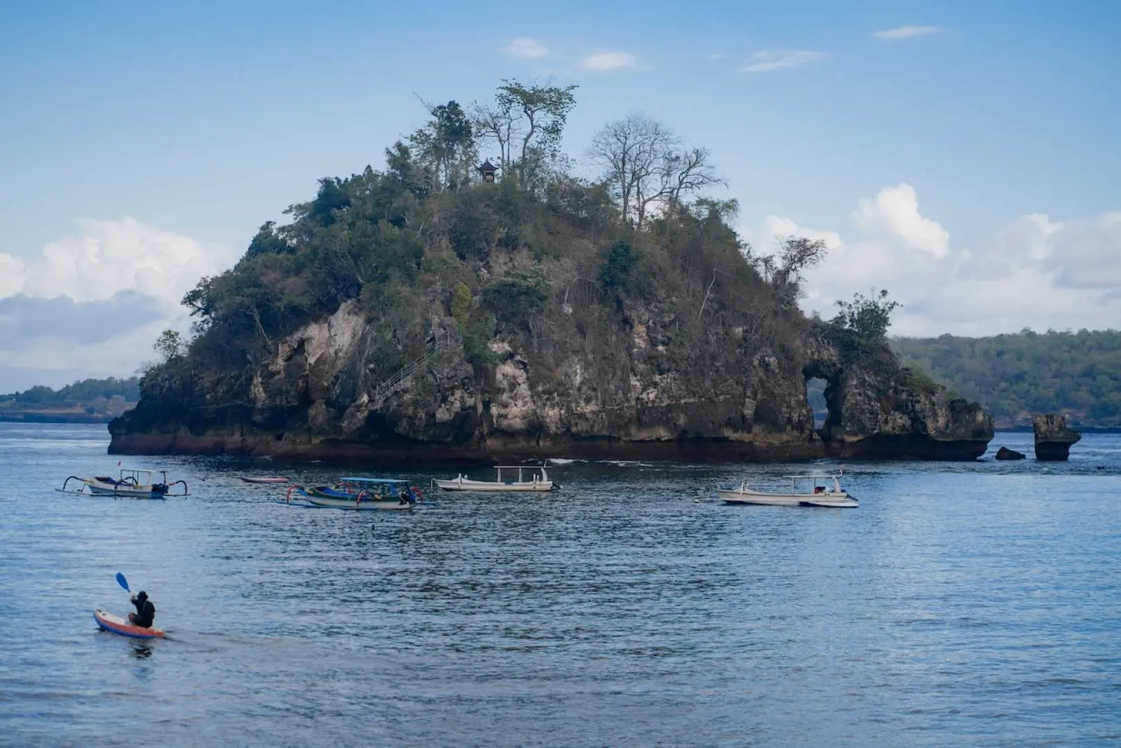 A lush green island and boats dot the calm waters around Nusa Penida, with a kayaker paddling, enjoying a peaceful water activity on a Bali day trip.