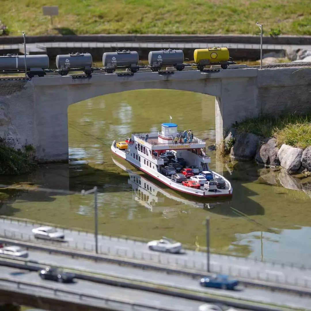 At Swissminiatur Lugano, a miniature train crosses a bridge, with boats sailing below and cars on the road, showcasing diverse Swiss transportation models.