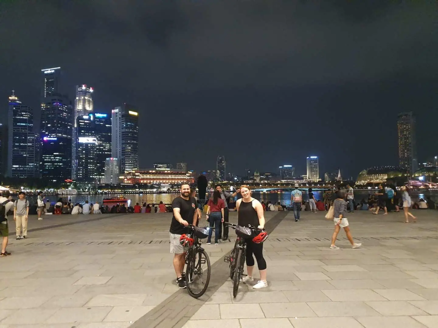 シンガポール・マリーナベイの夜景を背景に、自転車と一緒にポーズをとる2人の観光客。賑やかな都市の明かりが、ナイトサイクリングの楽しみを際立たせています。