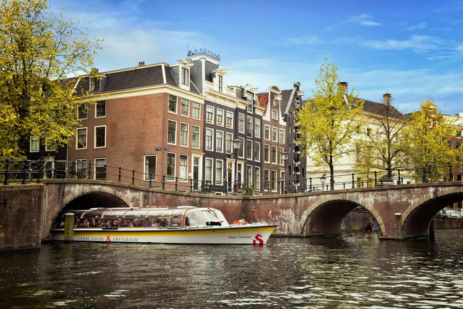 Amsterdam canal cruise boat under historic brick bridges, surrounded by traditional Dutch canal houses. Pass activity.