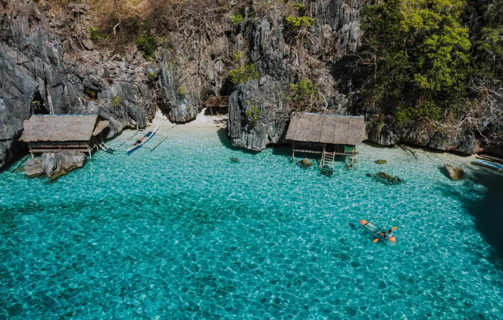 An aerial view of a traveler snorkeling in the crystal-clear turquoise waters of a secluded Coron lagoon, surrounded by majestic limestone cliffs and rustic huts.
