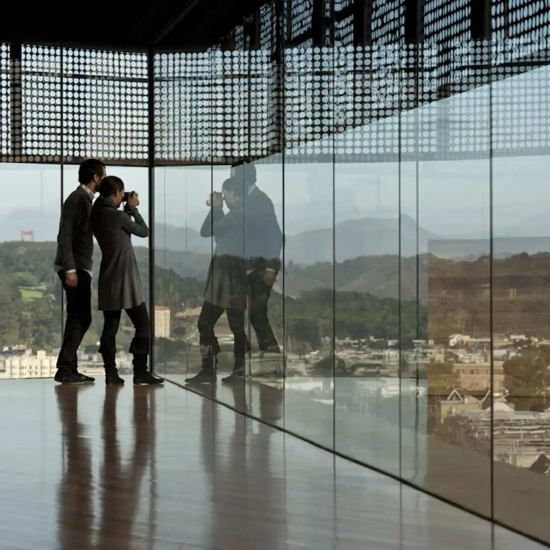 Visitors inside San Francisco's de Young Museum observation tower, enjoying panoramic views of the city and Golden Gate Park through large glass windows, a unique sightseeing experience.