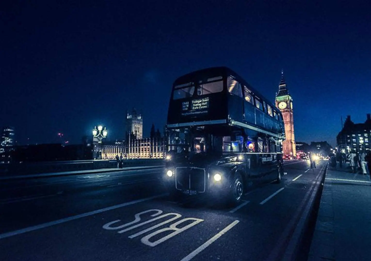 Classic black Routemaster Ghost Bus driving on a London street at night, with the iconic Big Ben sparkling in the background, signaling a unique night horror comedy tour experience.
