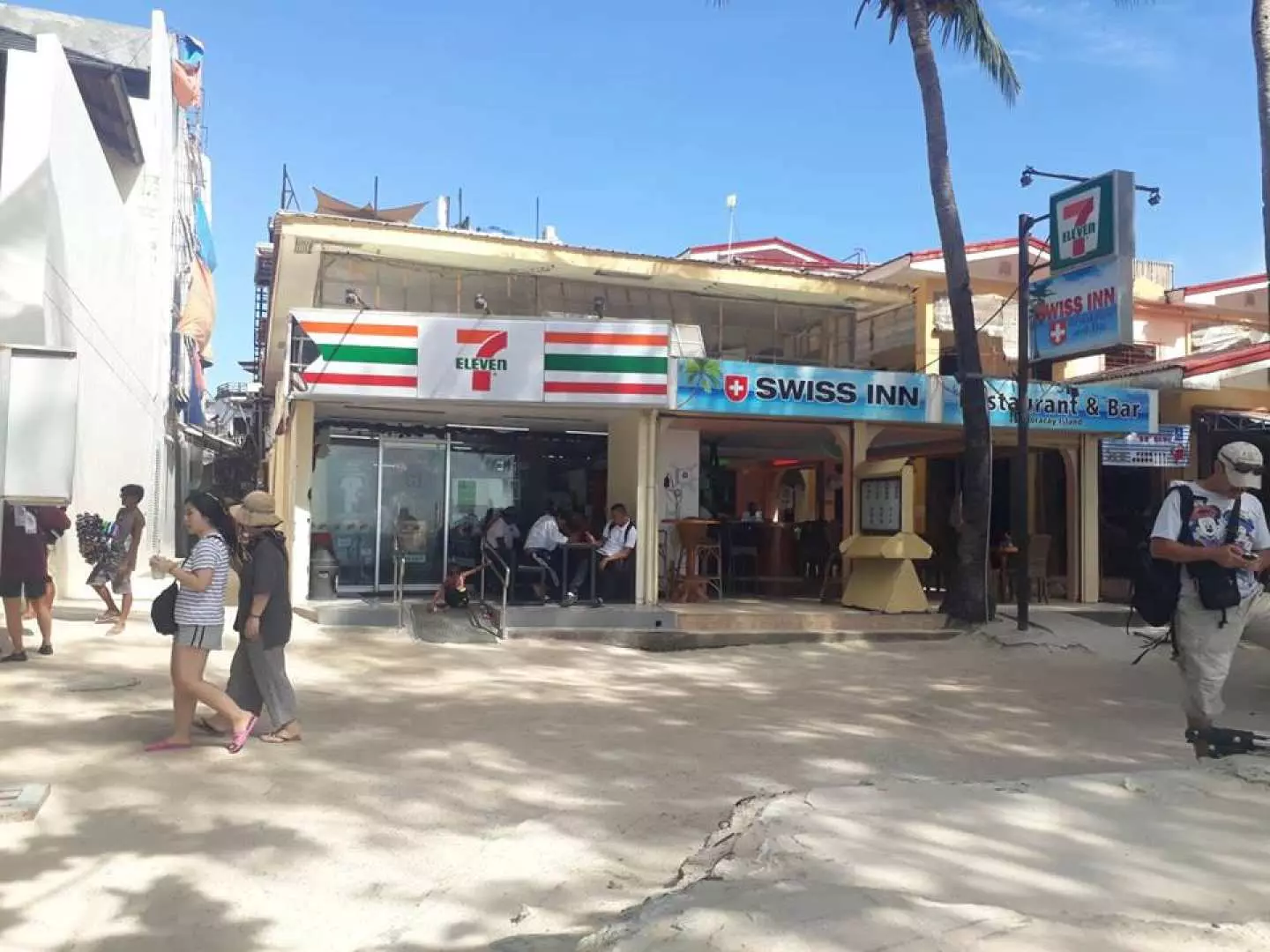 ภาพทิวทัศน์ริมหาดในโบราไกย์ แสดงร้านสะดวกซื้อ 7-Eleven และร้านอาหาร Swiss Inn ซึ่งมักใช้เป็นจุดนัดพบสำหรับกิจกรรมและบริการในท้องถิ่น