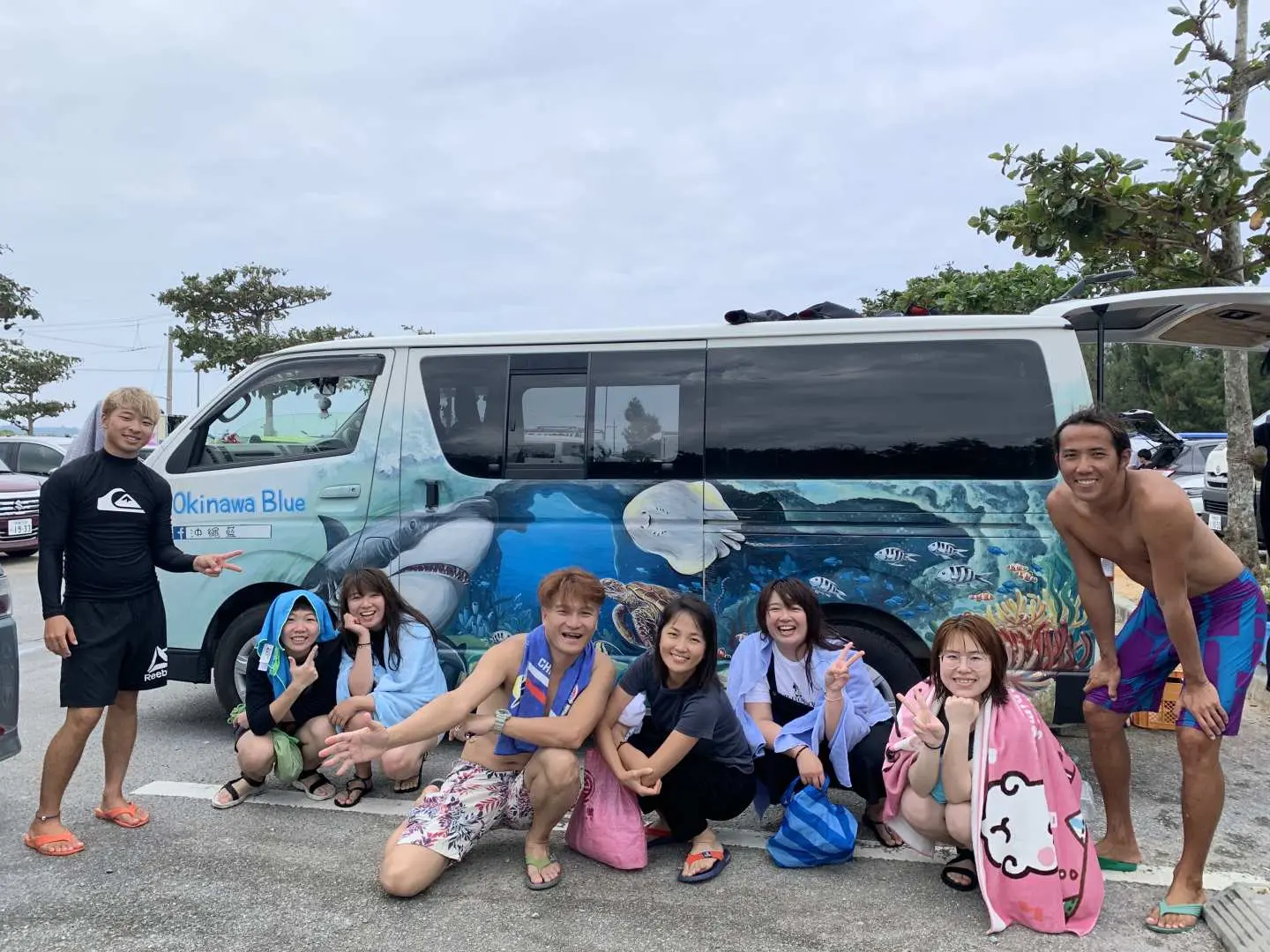 A cheerful group of participants and instructors gathered for a photo in front of the 'Okinawa Blue' shuttle, celebrating a day of Blue Cave and water activities.