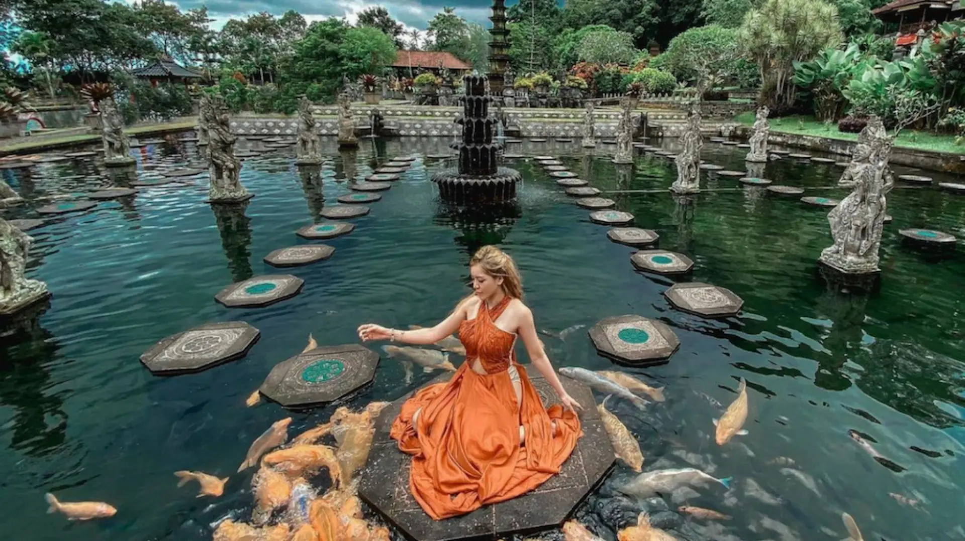 A woman in a vibrant orange dress sits gracefully on a central stepping stone in Bali's Tirta Gangga Royal Water Palace, surrounded by a multitude of colorful koi fish eager for interaction.