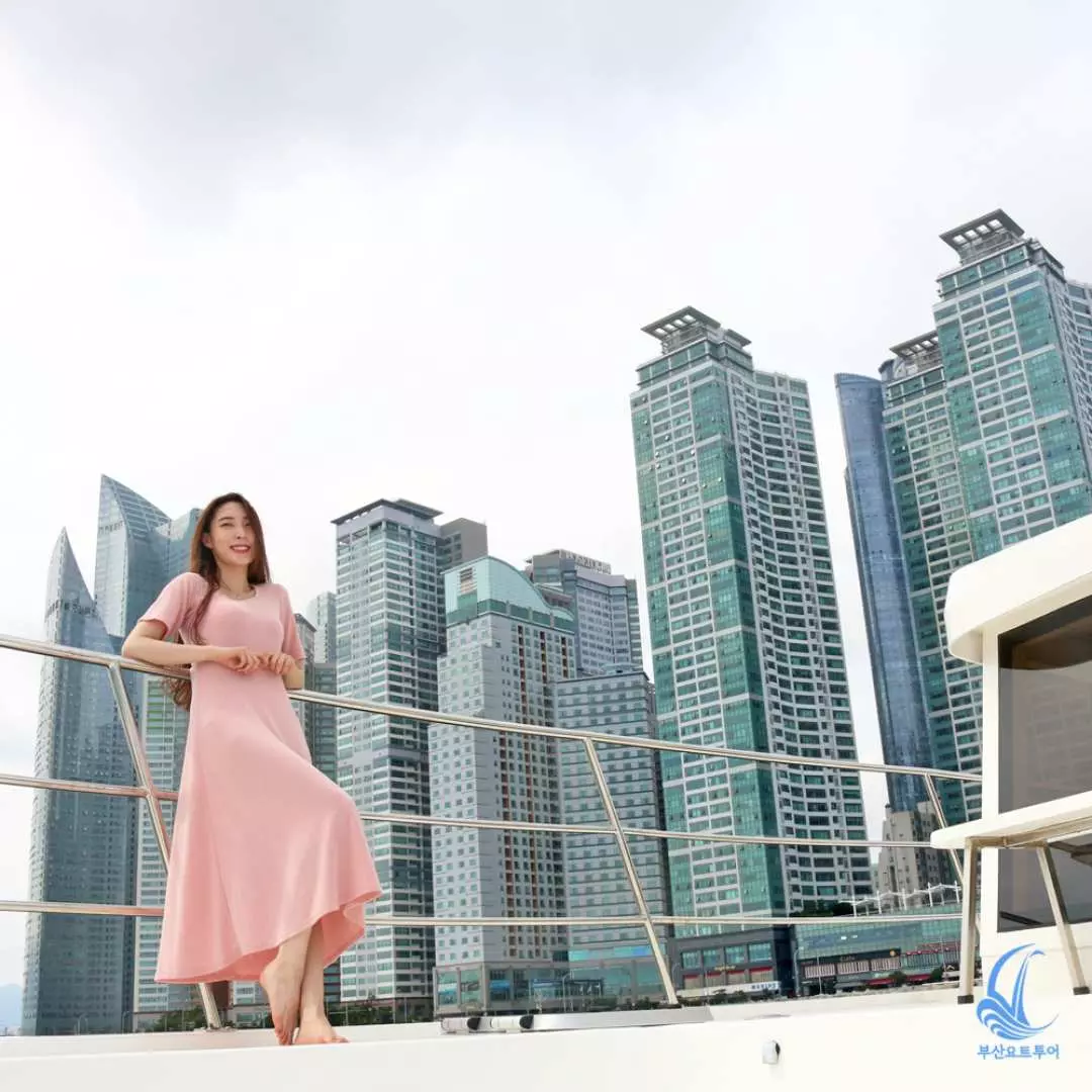 Một phụ nữ mặc váy dài màu hồng duyên dáng tựa vào lan can du thuyền sang trọng ở Busan, với cảnh biển ngoạn mục và đường chân trời thành phố đầy các tòa nhà cao tầng ở phía sau.