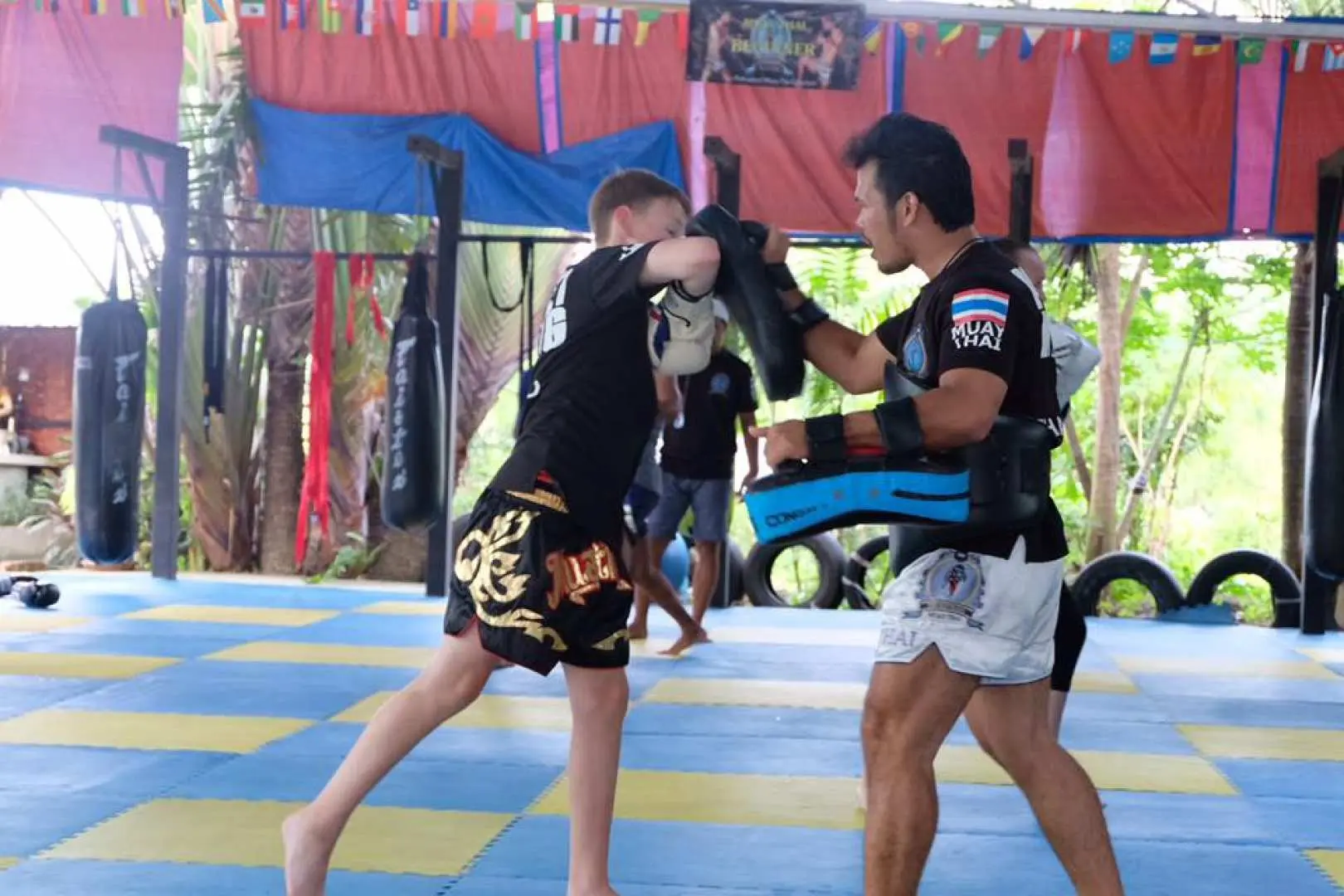 In a Muay Thai class in Kathu, Phuket, a young student in traditional Muay Thai shorts practices knee strikes under instructor guidance, demonstrating fundamental Muay Thai skills.