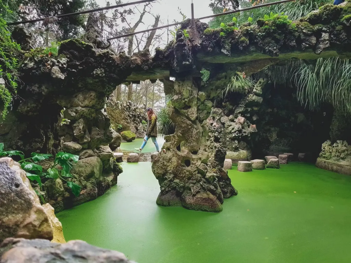 A visitor at Quinta da Regaleira in Sintra, Portugal, navigating stone bridges and stepping stones over a vibrant green lake, experiencing the unique joy of exploring the estate's mysterious gardens.