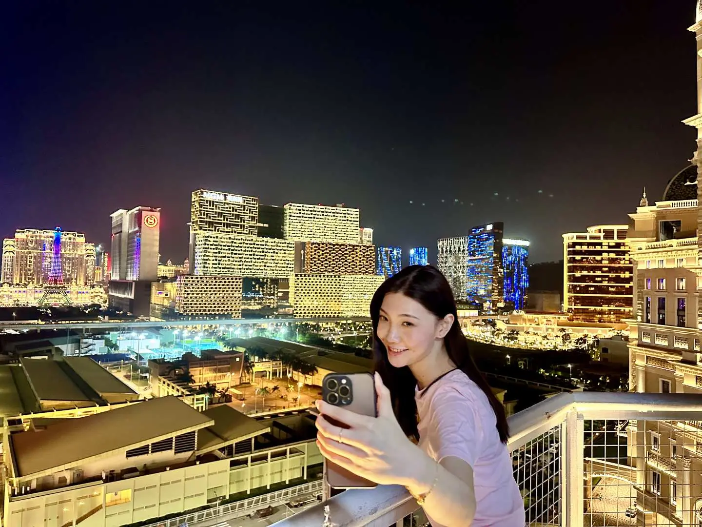 マカオZIPCITYタワーの夜間展望台で、若い女性がきらめくマカオの夜景を背景に自撮り。忘れられない夜の高所体験を楽しんでいる。