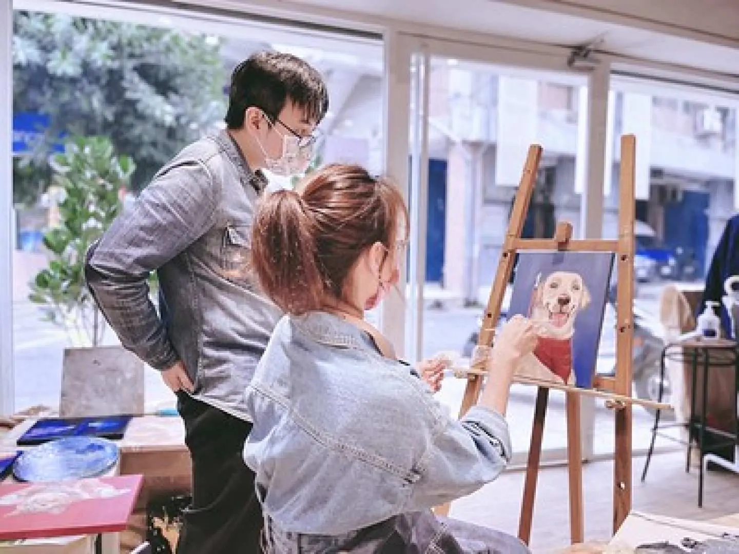 Sanchong oil painting: couple in bright studio. Woman paints vivid pet portrait, man watches, enjoying relaxed art.