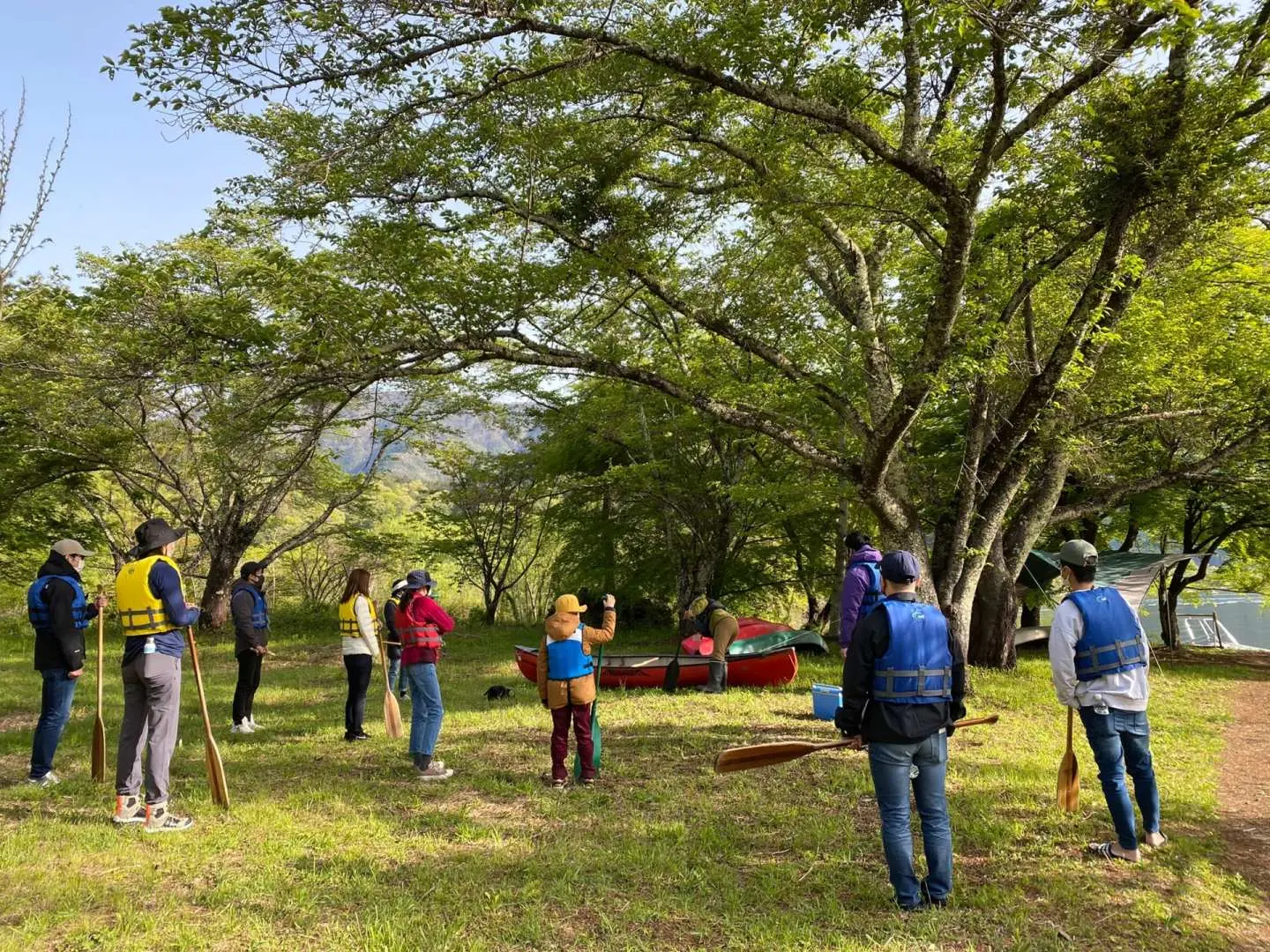 A group of canoe participants listens attentively to their guide under the shade of lush green trees on the grassy shore, with canoes pulled ashore. This scene captures the essential pre-activity safety briefing for the 120-minute Lake Kawaguchiko canoe course.