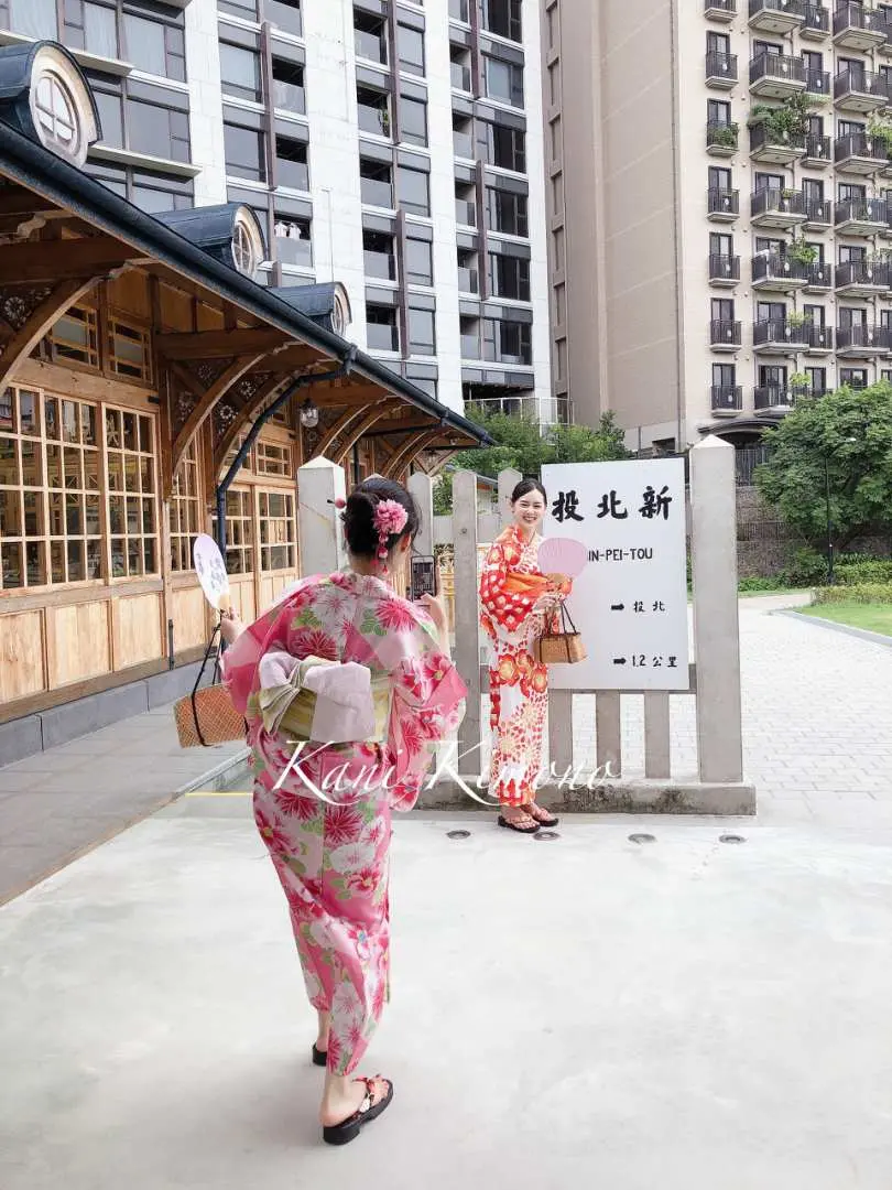 兩位女性穿著鮮豔浴衣，在北投「新北投」地標前合影留念，手持扇子與日式小包，體驗日式街景穿梭樂趣。