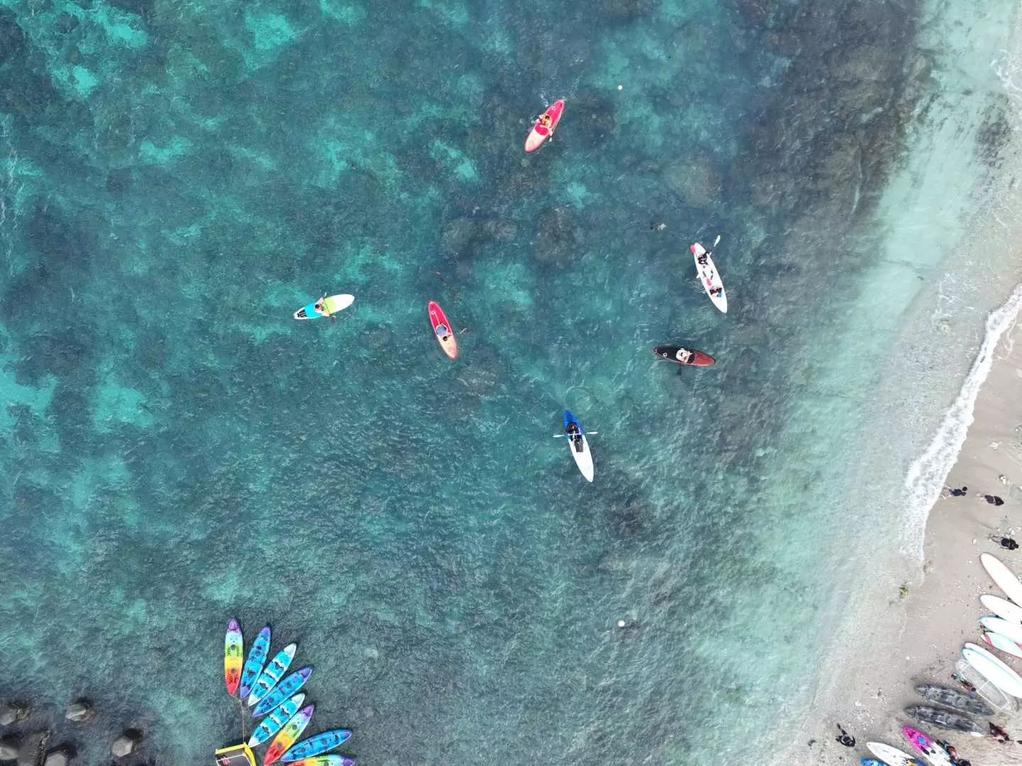 Aerial view of colorful SUP stand-up paddleboards floating on the clear coral reef waters of Xiaoliuqiu, Taiwan, with tourists enjoying the water activity and more boards on the sandy shore.