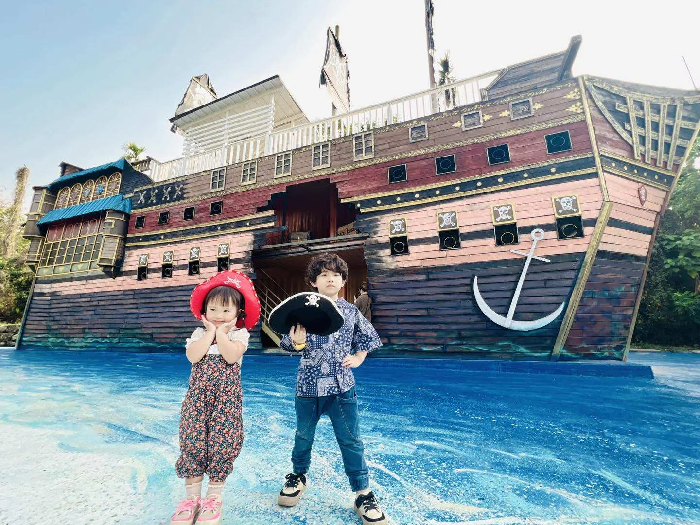 Two children pose in front of the colossal real-life pirate ship and blue ocean-themed landscape at Nantou Pirate Village, offering a whimsical family travel experience.