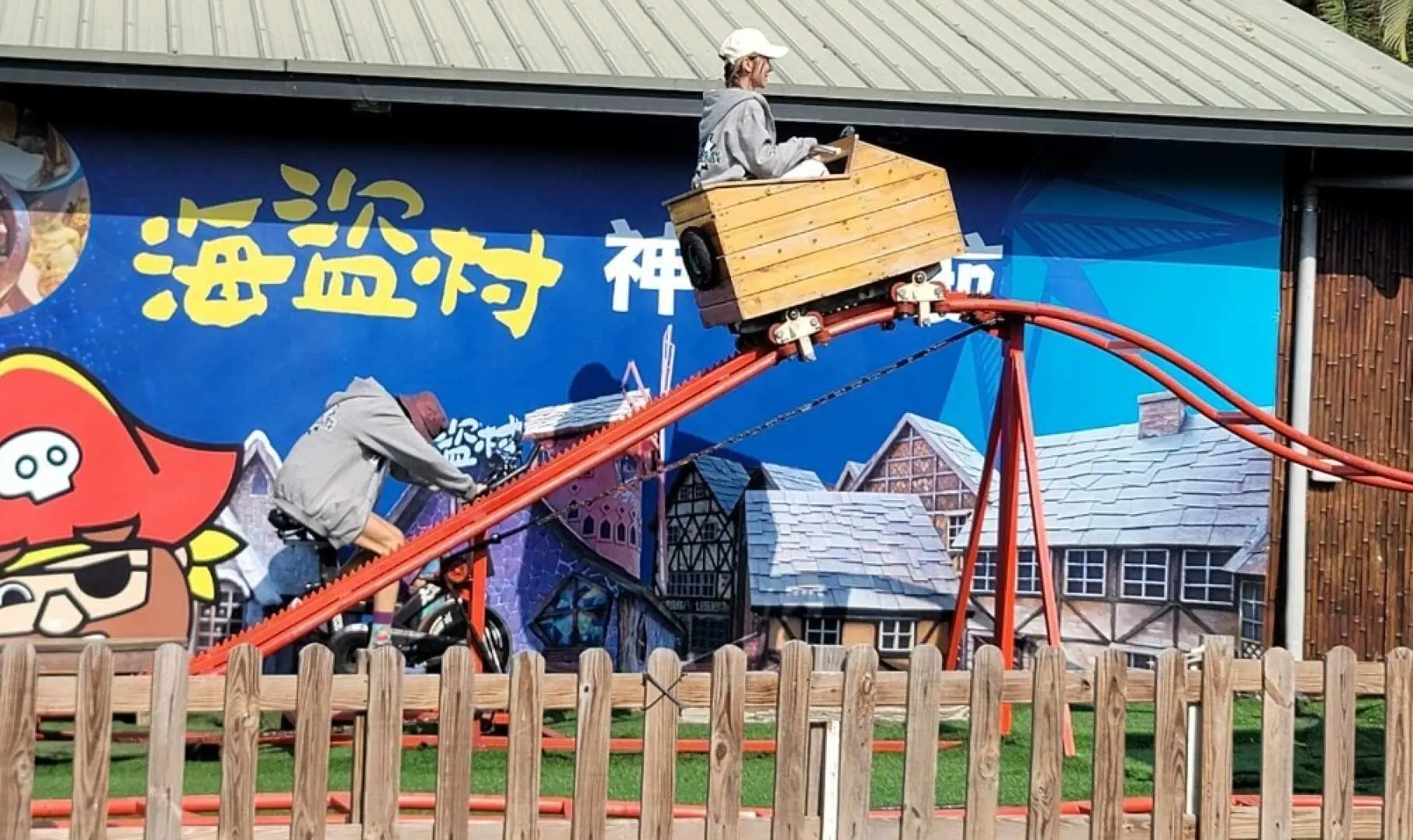 A child enjoying the 'God-Devil Ride' mini rollercoaster at Nantou Pirate Village, riding solo in a wooden cart on a colorful track, experiencing outdoor amusement.