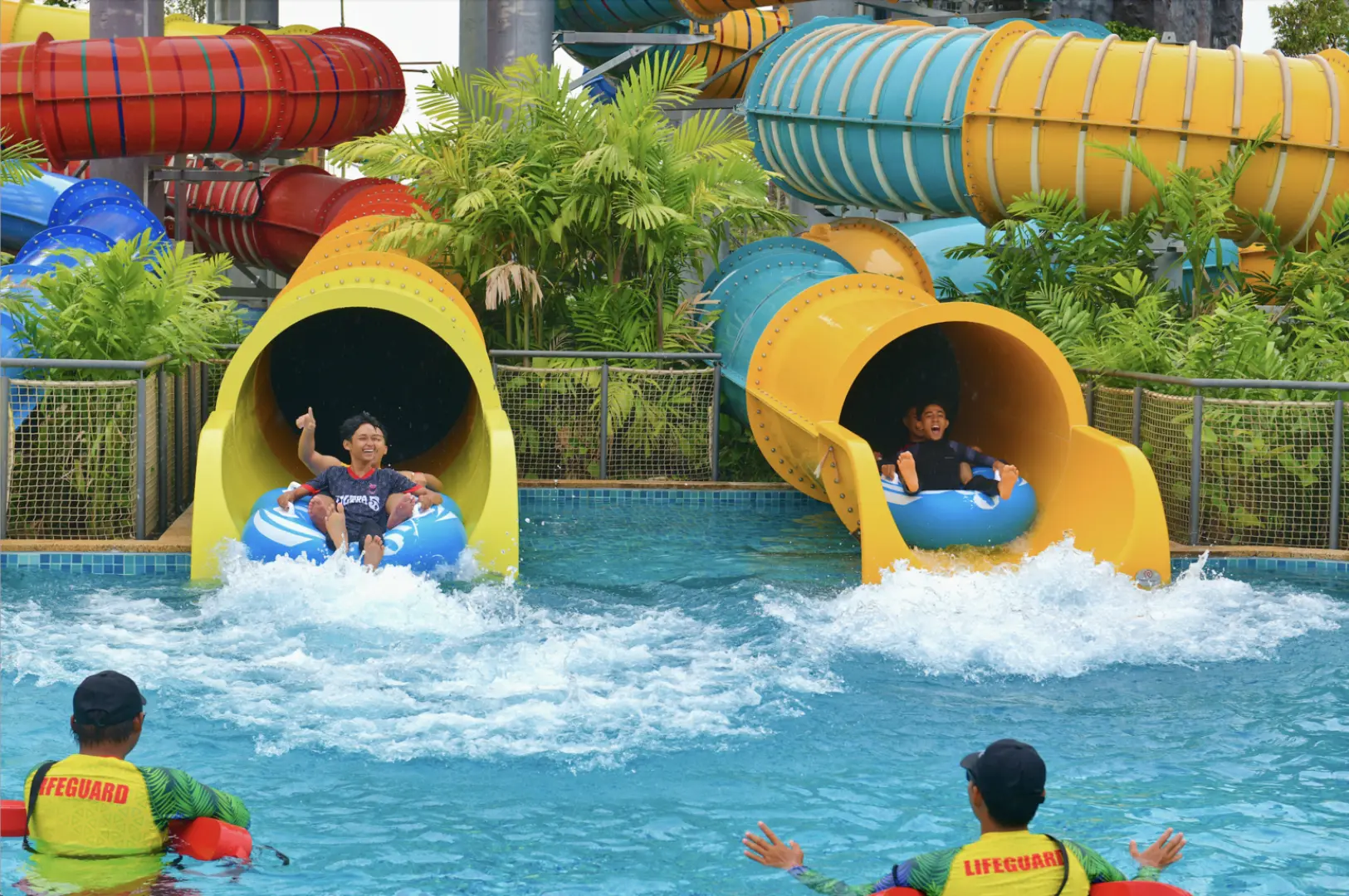 Two children excitedly riding inflatable tubes down yellow water slides at SplashMania Water Park, splashing into the pool and enjoying family fun, with lifeguards ensuring safety.