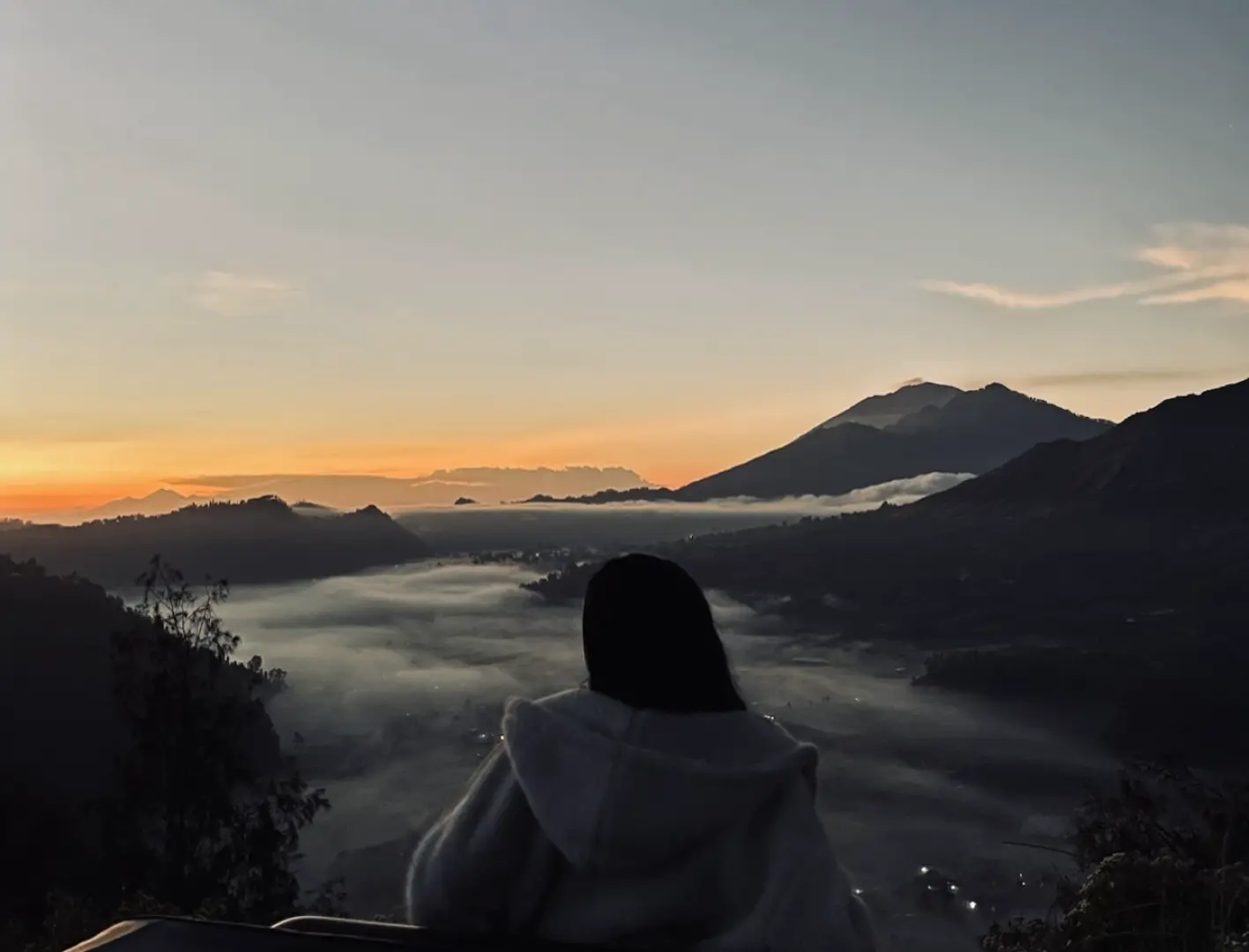 遊客背對鏡頭，在巴厘島巴圖爾山區欣賞日出美景，薄霧籠罩山谷，遠處山脈連綿，景色壯麗。