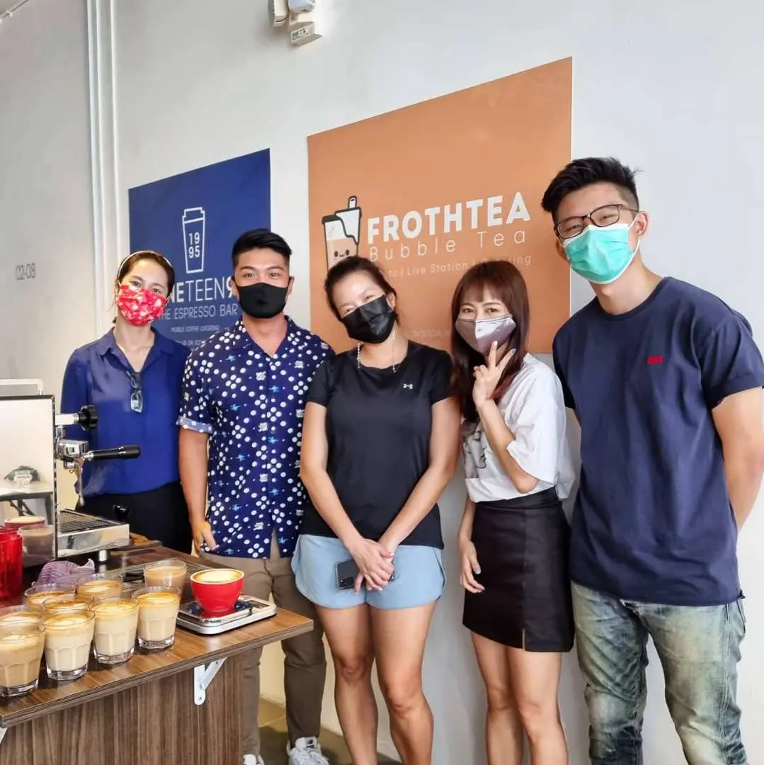 A group of five smiling participants and instructors, wearing masks, pose happily in front of the Nineteen95 branding, showcasing various coffee drinks and a joyful learning atmosphere at the Singapore workshop.