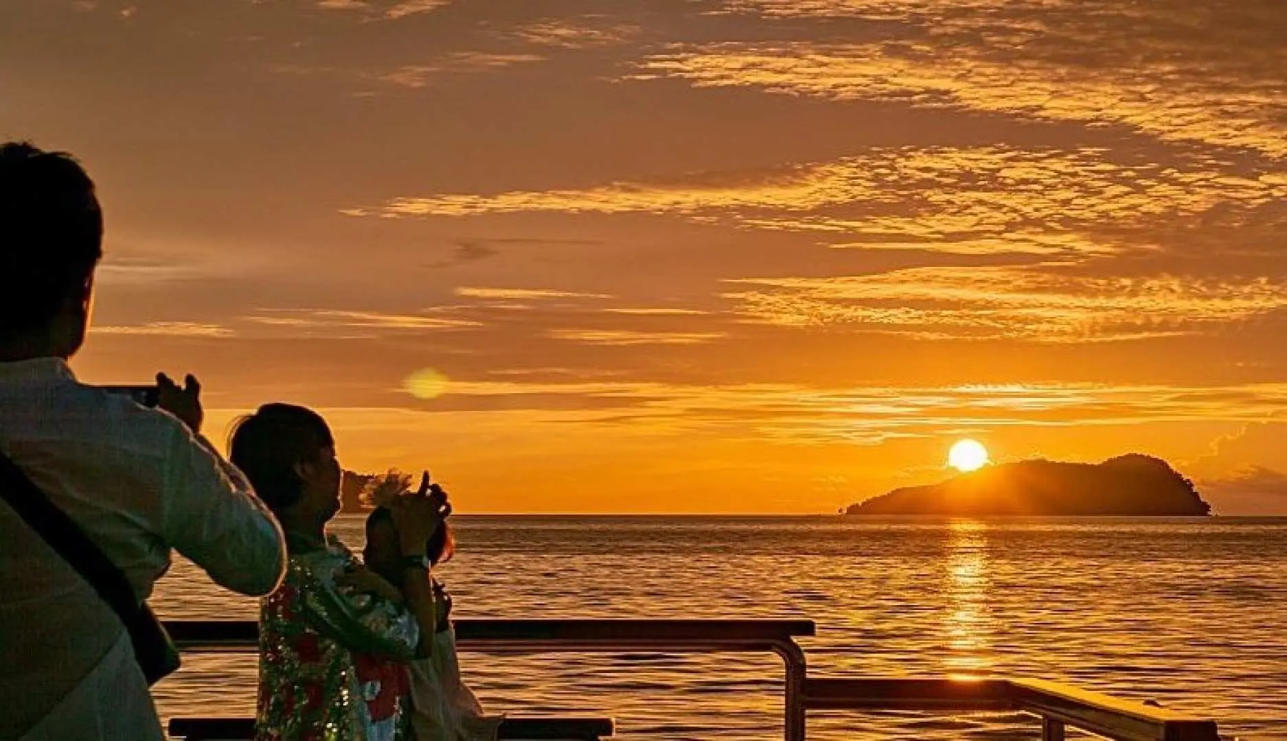 A parent and child on the Kota Kinabalu sunset cruise deck, admiring the magnificent orange sunset over the South China Sea, featuring a distant island silhouette.