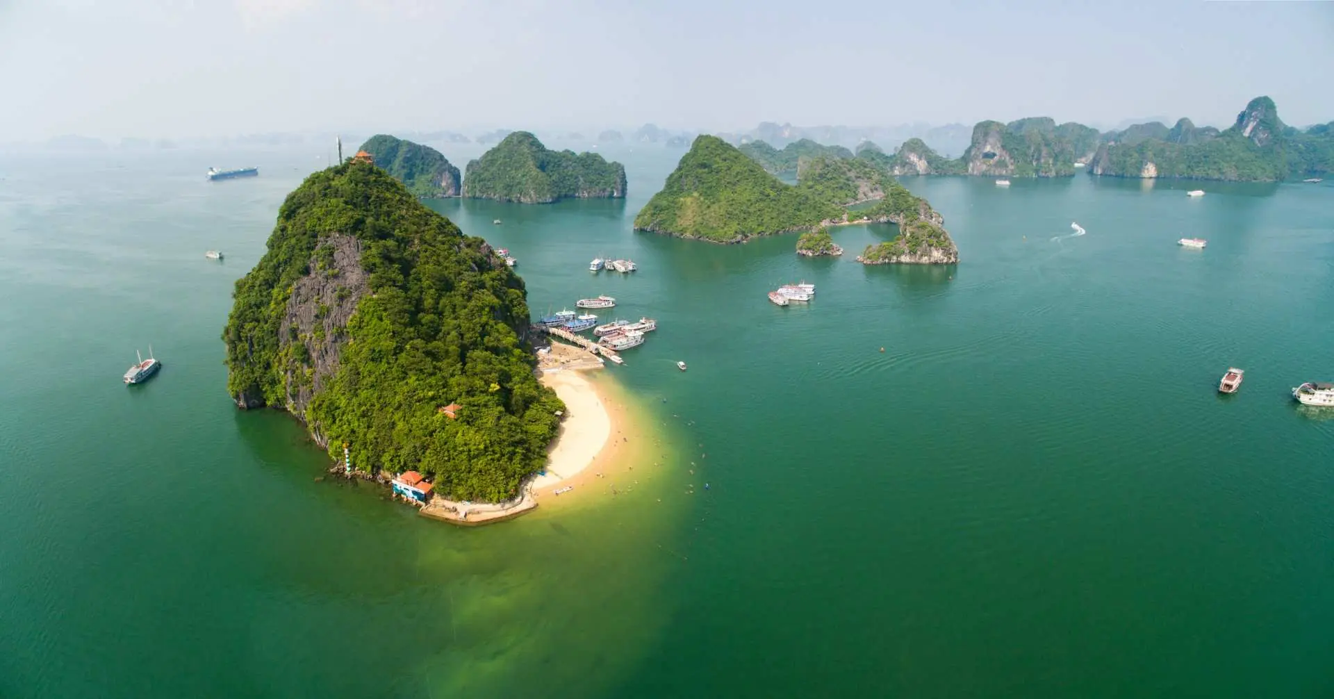 Aerial view of Ti Top Island in Halong Bay, featuring its pristine sandy beach and lush green conical peak, surrounded by turquoise waters with boats anchored.