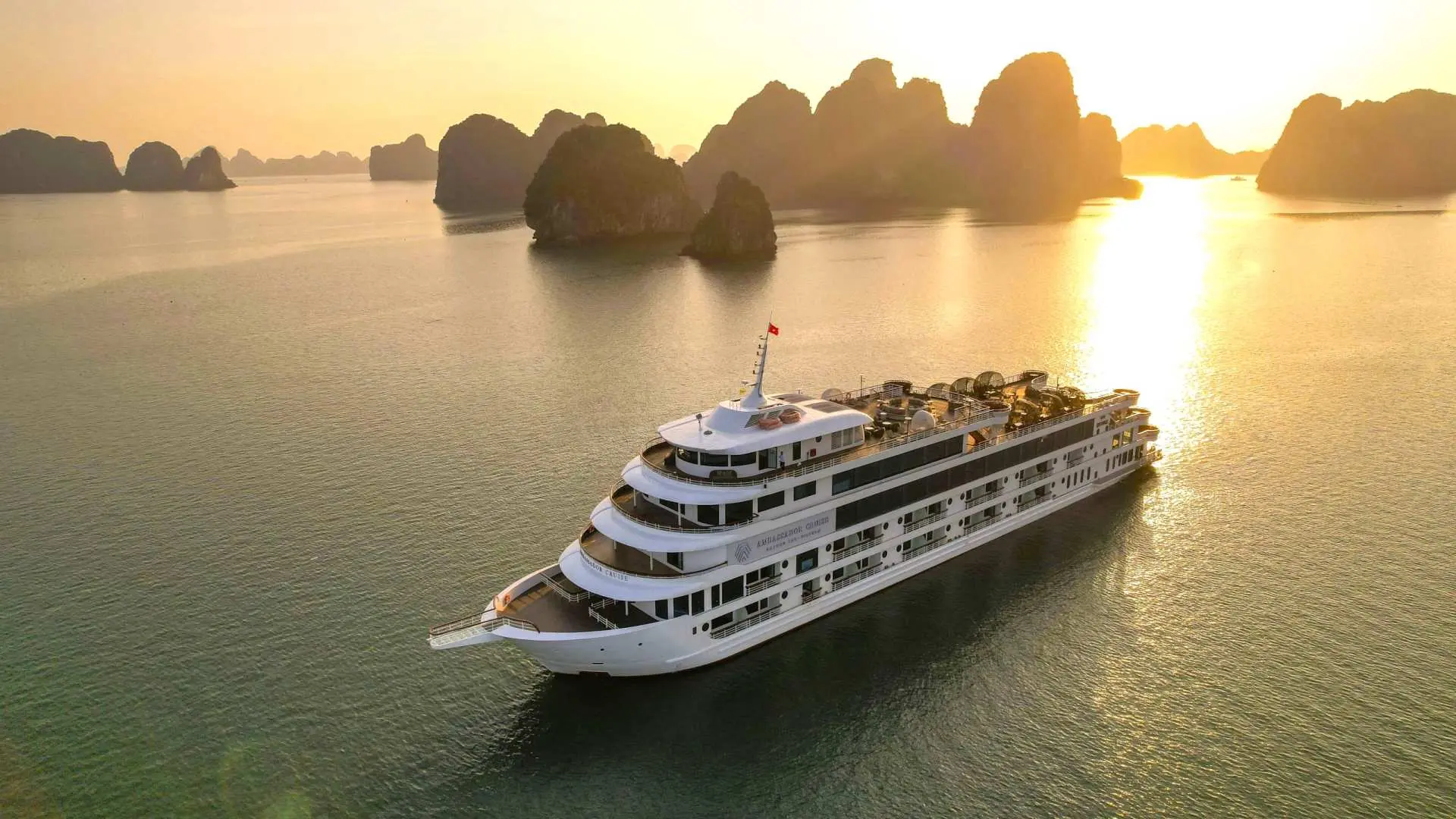 The Ambassador luxury cruise majestically sails through Halong Bay at sunset, with golden light illuminating the iconic limestone karsts and calm waters.