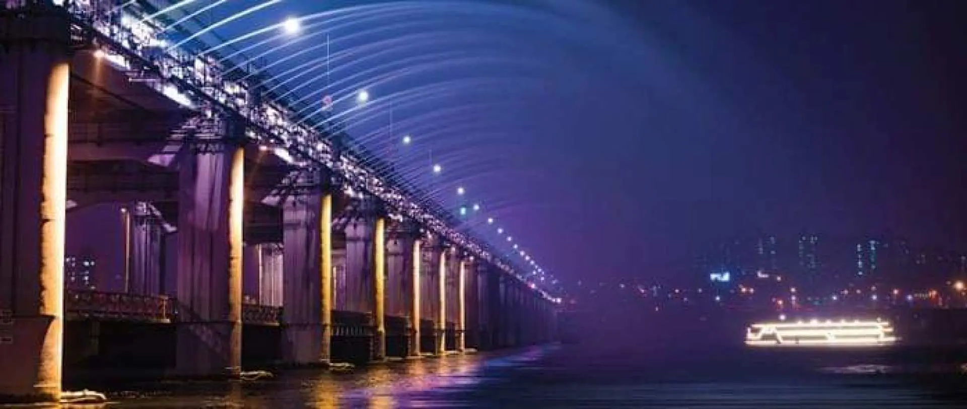韓國首爾盤浦大橋的璀璨夜景，橋身被紫色與黃色燈光點亮，漢江上可見遊船，氣氛浪漫。