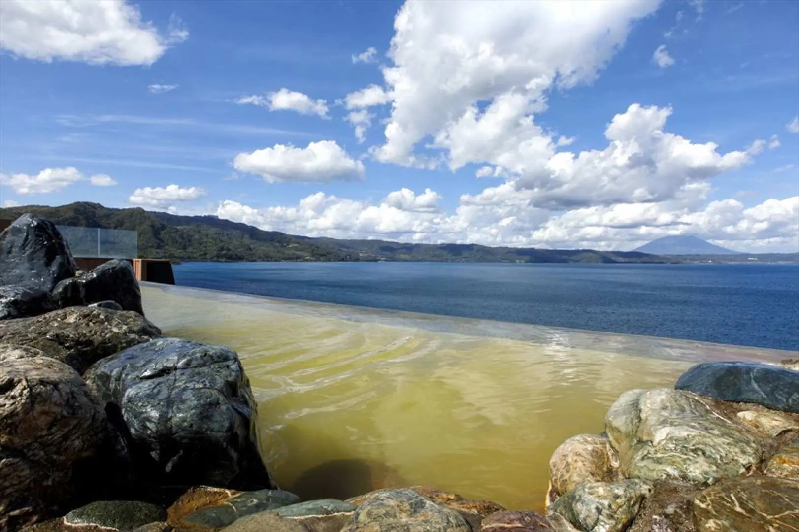 Spectacular open-air onsen by Lake Toya in Hokkaido, featuring unique yellowish-green waters that blend seamlessly with the azure lake and distant mountains, offering ultimate relaxation.