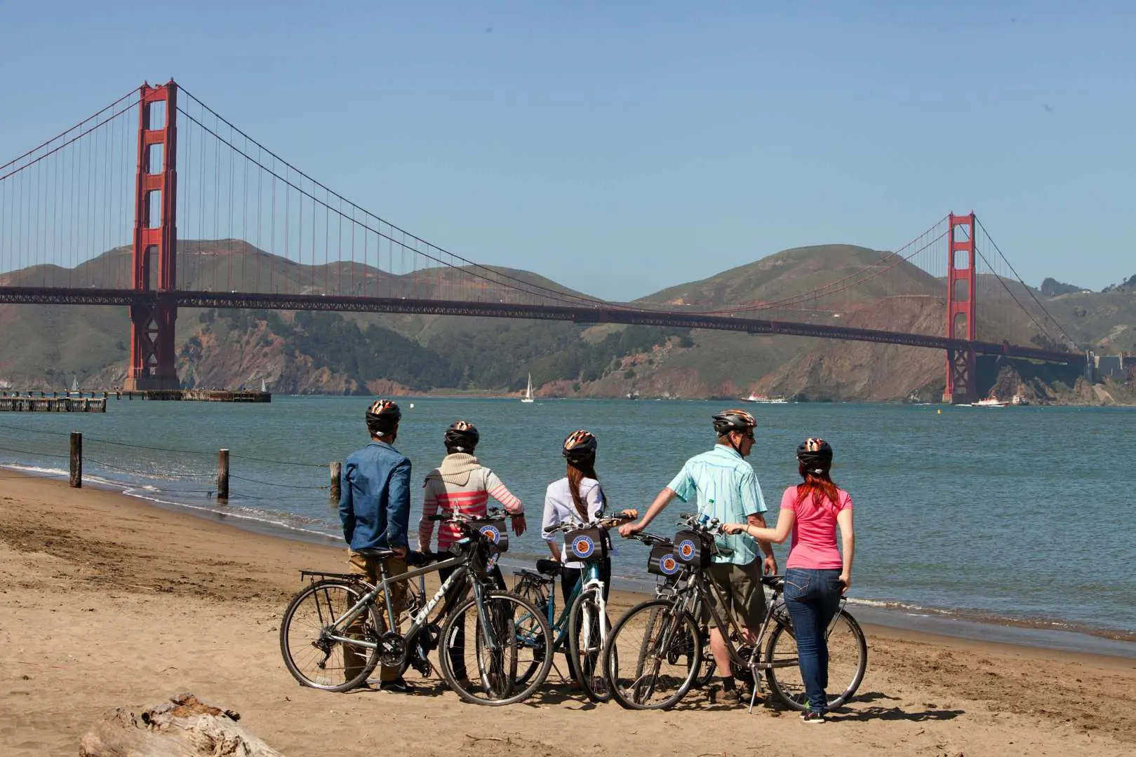 Visitors pause on a San Francisco beach with their bikes to admire the majestic Golden Gate Bridge in the distance, enjoying coastal views during their bike tour.