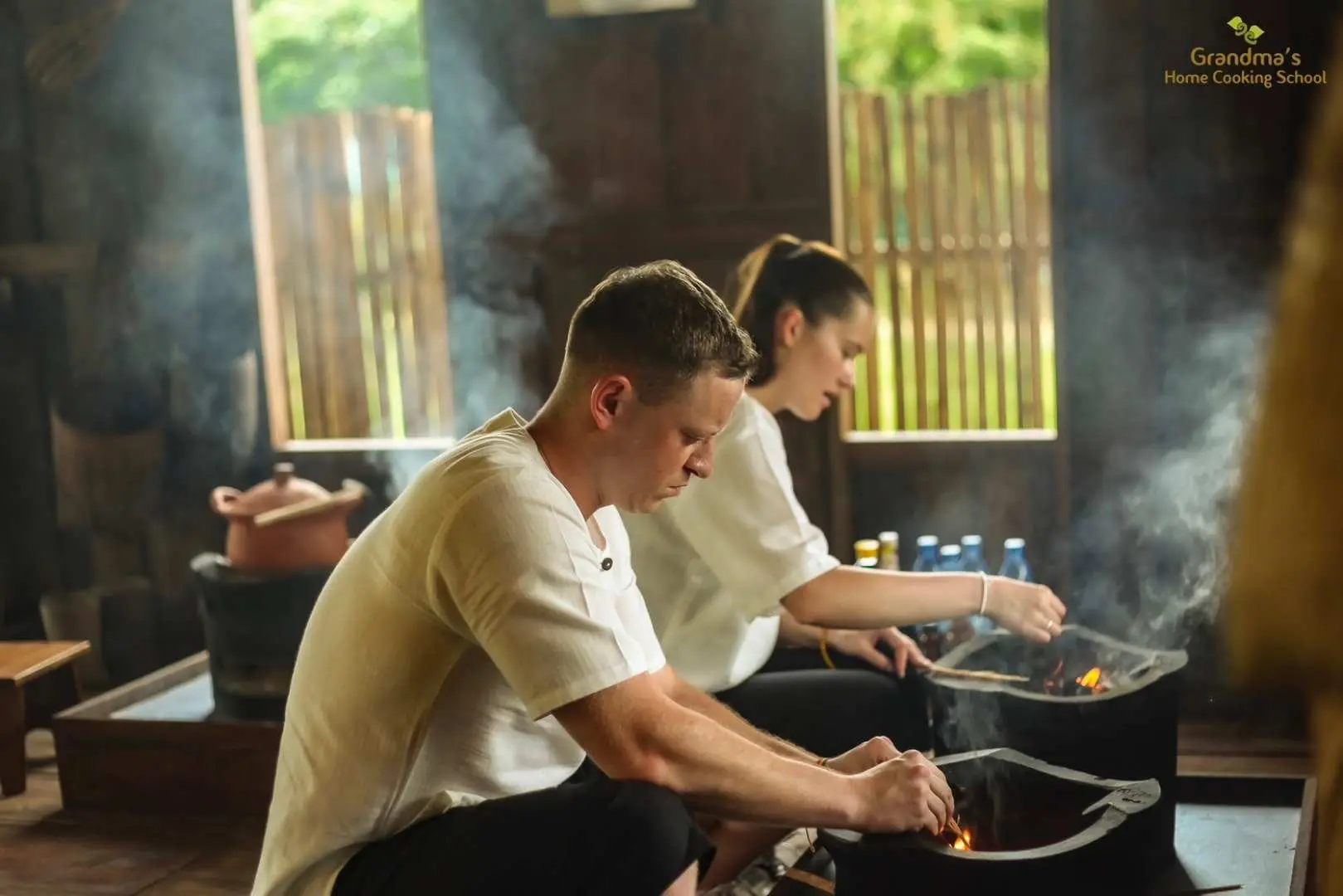 Two students in a Chiang Mai cooking class diligently prepare Northern Thai dishes on traditional charcoal stoves, with steam rising in the rustic wooden kitchen.