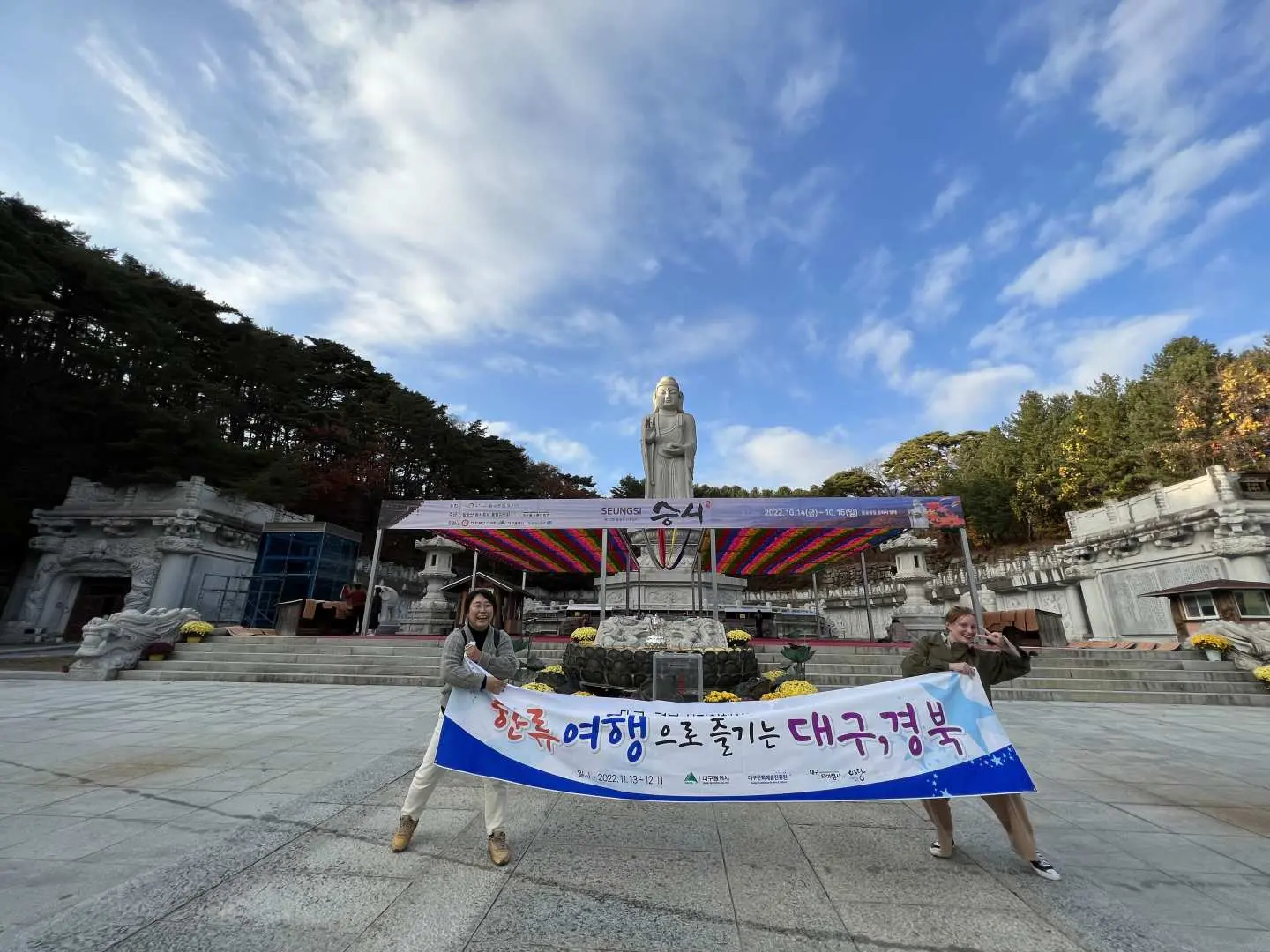 Two visitors holding a 'Enjoy Daegu, Gyeongbuk with Korean travel' banner in front of a majestic Buddha statue and solemn Buddhist architecture at Palgongsan, Daegu.