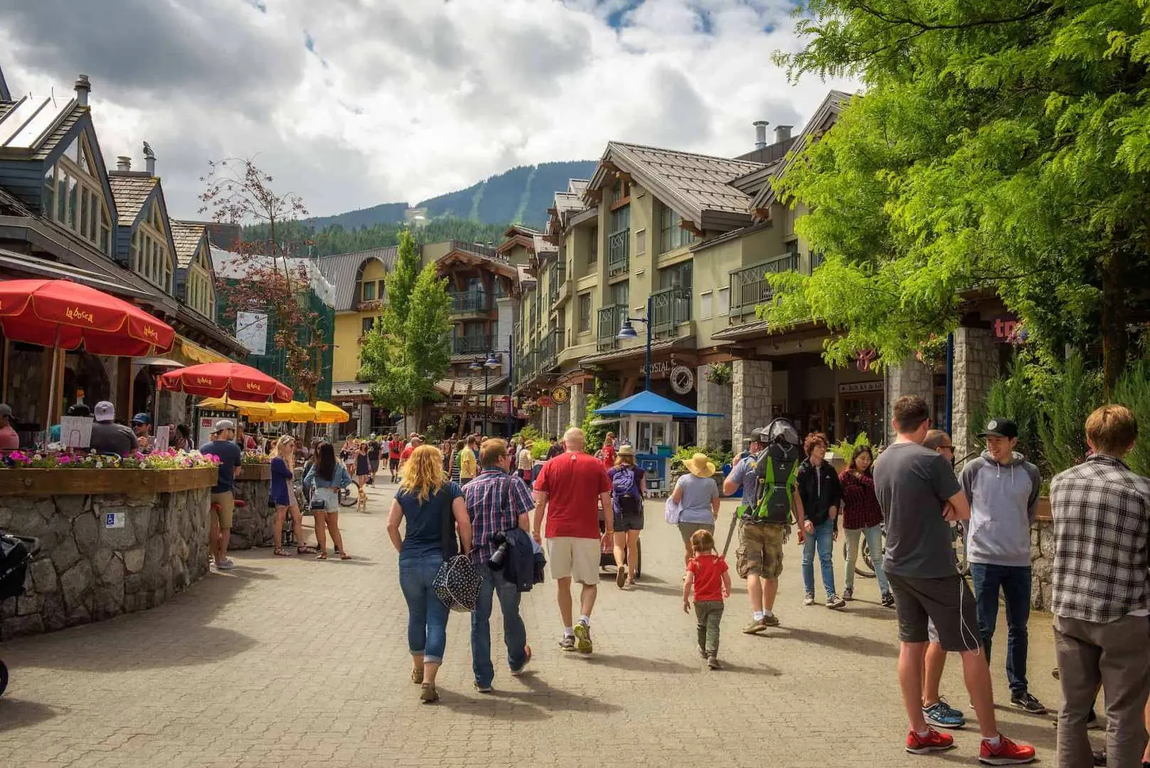A lively pedestrian street in Whistler Village, Canada, bustling with visitors strolling, shopping, and dining at outdoor cafes, surrounded by charming alpine-style buildings and lush greenery, immersing in local culture.