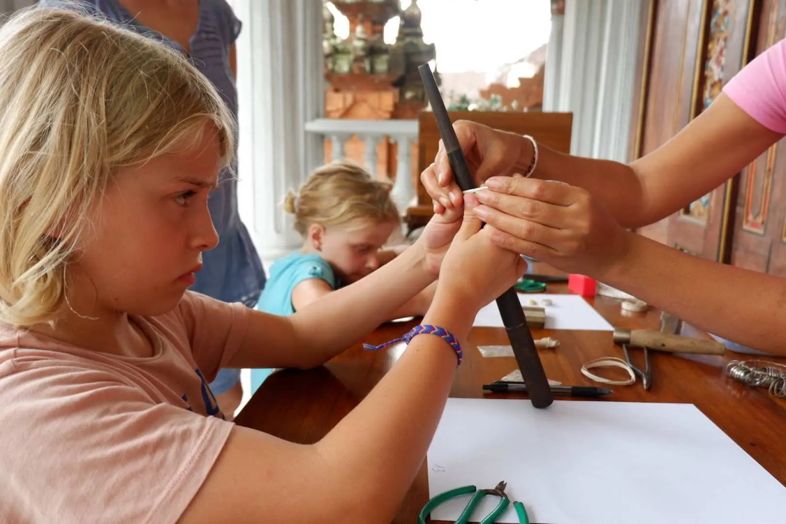 In a child-friendly Bali silver workshop, a young student is gently guided by an adult's hands to use tools for crafting their own silver jewelry, fostering artistic interest.
