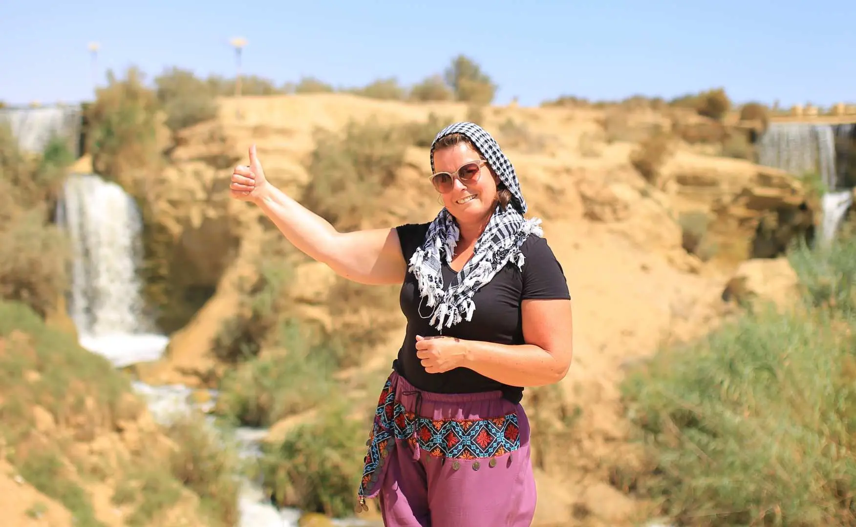 A happy female traveler giving a thumbs-up in front of the picturesque Wadi El Rayan waterfalls, showcasing the stunning natural beauty of the Egyptian desert landscape.