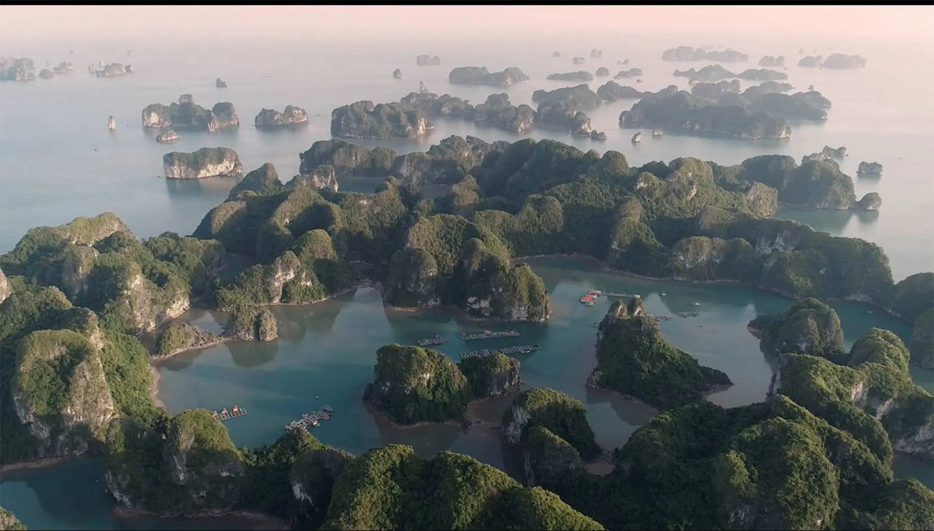 An expansive aerial view of Halong Bay at dusk or dawn, showcasing numerous limestone islands and calm waters, with visible fishing villages adding to the serene bay atmosphere.