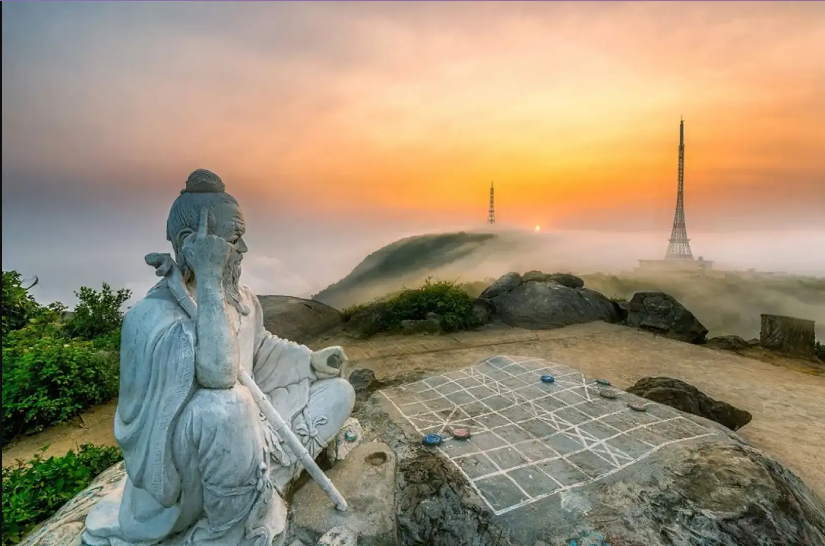 A serene stone statue of an ancient figure seated by a chessboard, overlooking a mystical, mist-shrouded Son Tra Mountain landscape in Da Nang at dawn or dusk, near the Linh Ung Pagoda.